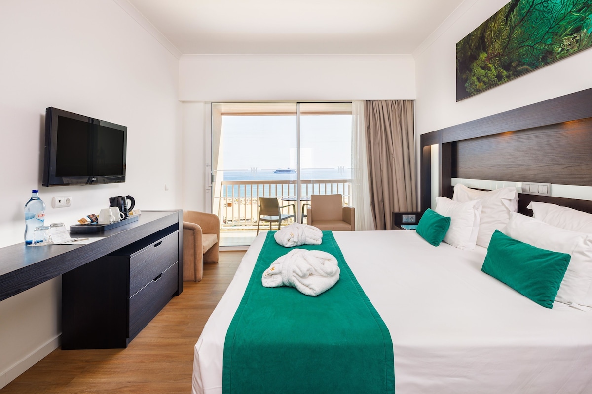 A hotel room with a bed, a desk, a TV, a balcony with a view, and a painting on the wall.