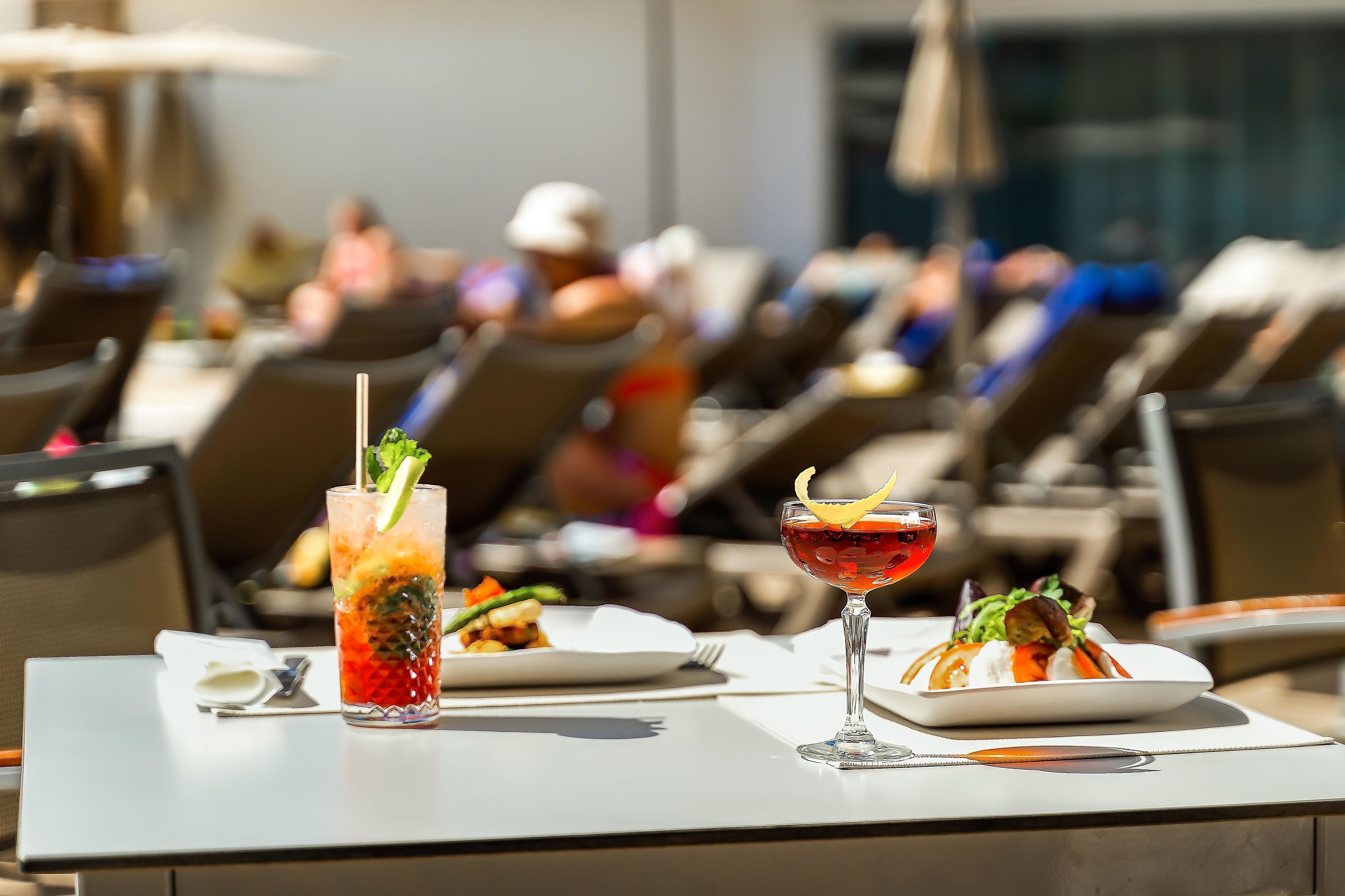 A dining table with two plates of food and two drinks, set on an outdoor terrace with sun loungers in the background.