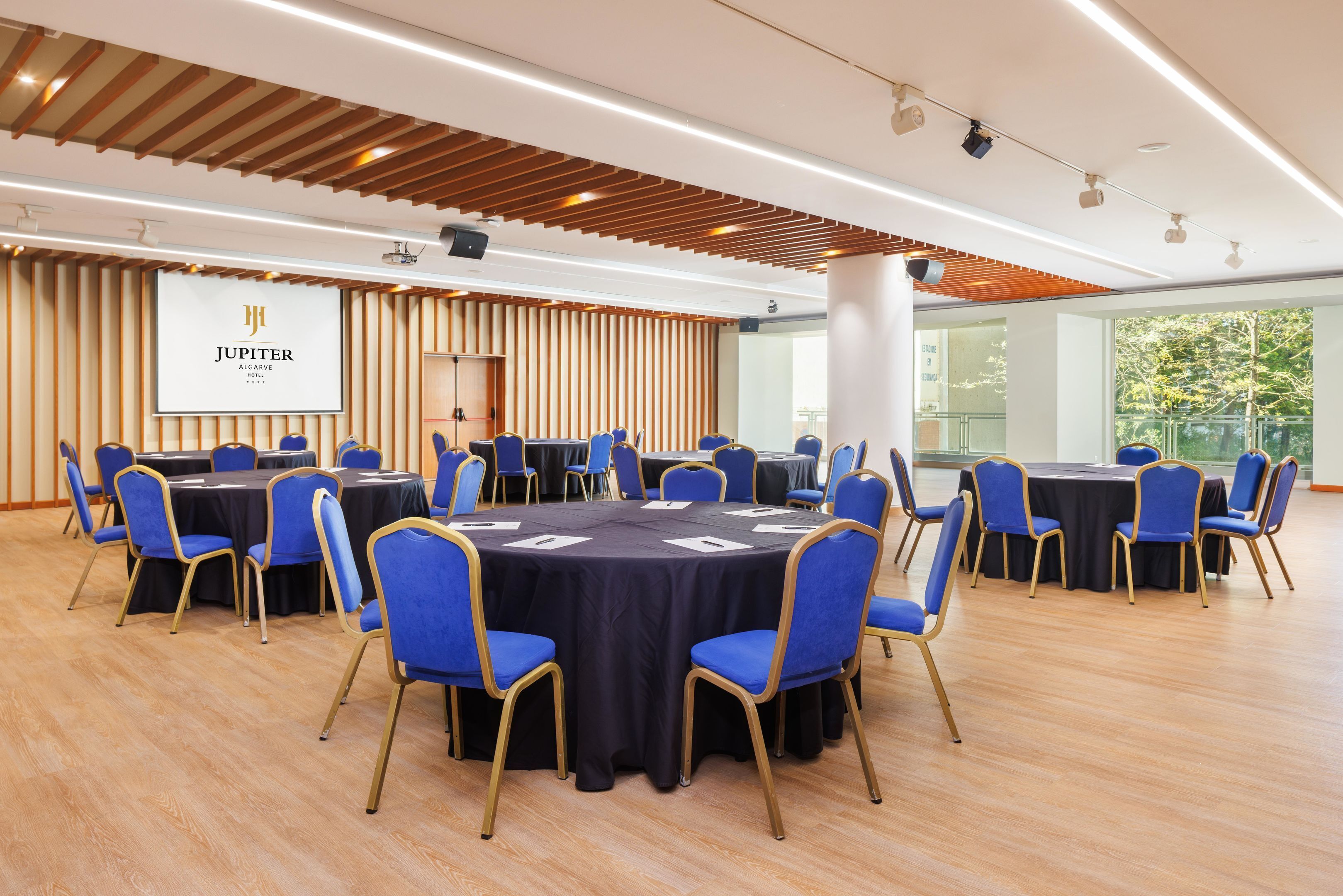 A conference room with round tables, blue chairs, and a large screen displaying 'JUPITER ALGARVE'.