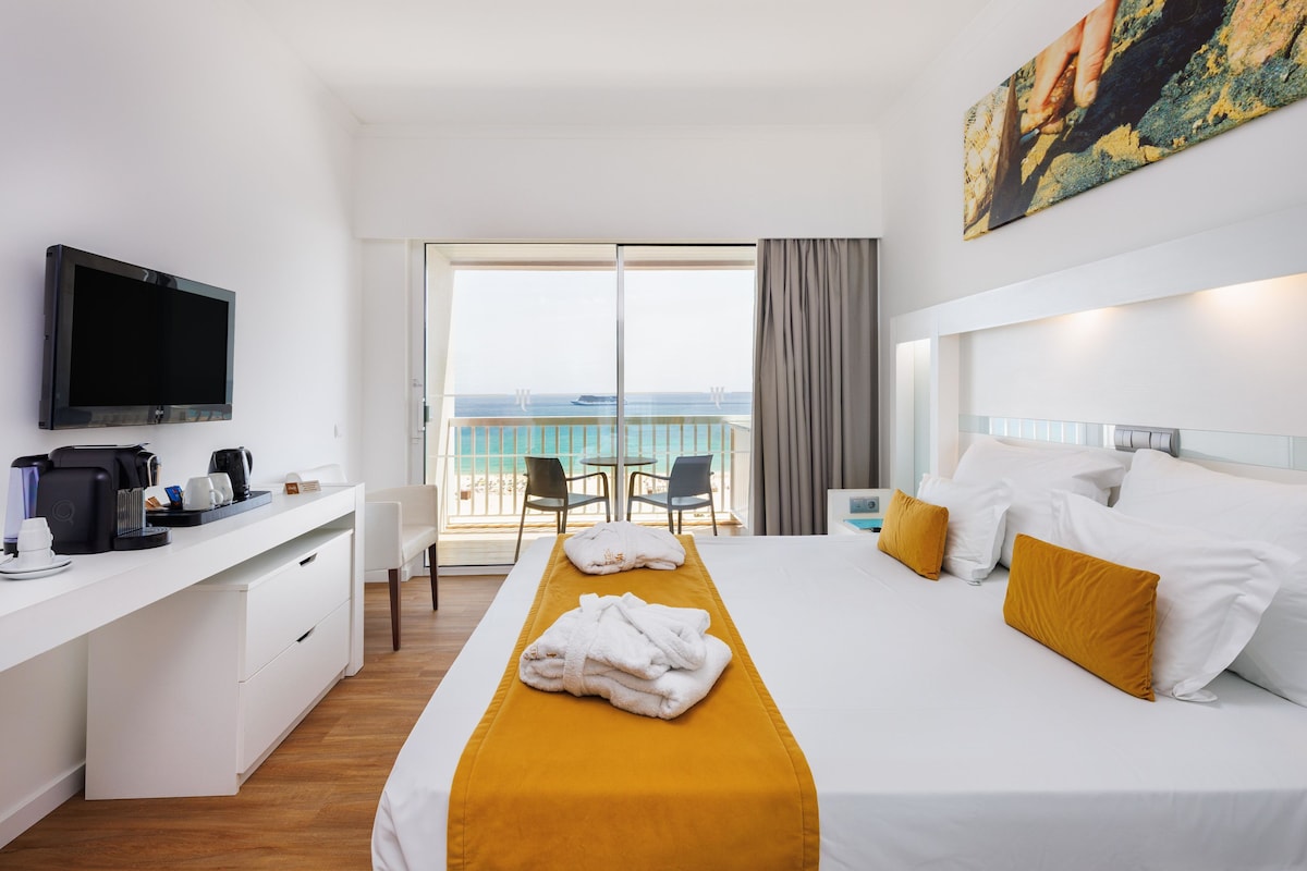A hotel room with a bed, a desk with a TV, a coffee maker, and a view of the sea through large windows.