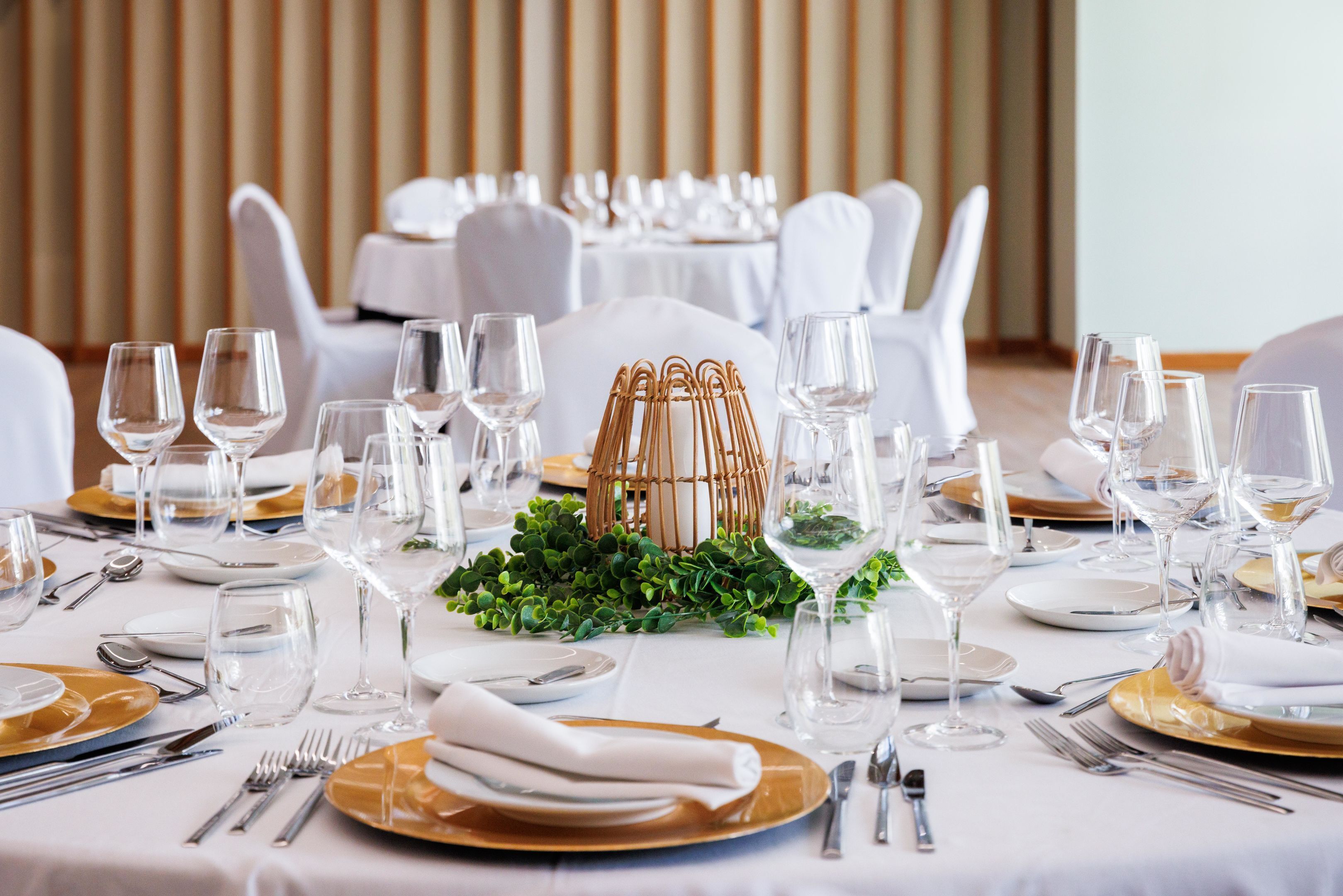 A dining table set with white and gold plates, silverware, and clear wine glasses, centered with a decorative wicker basket and greenery.