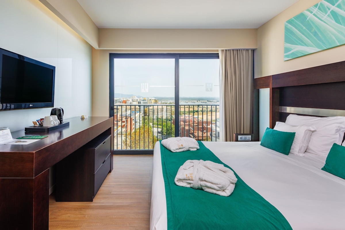A hotel room with a large bed, a desk with a flat-screen TV, and a balcony with a city view.