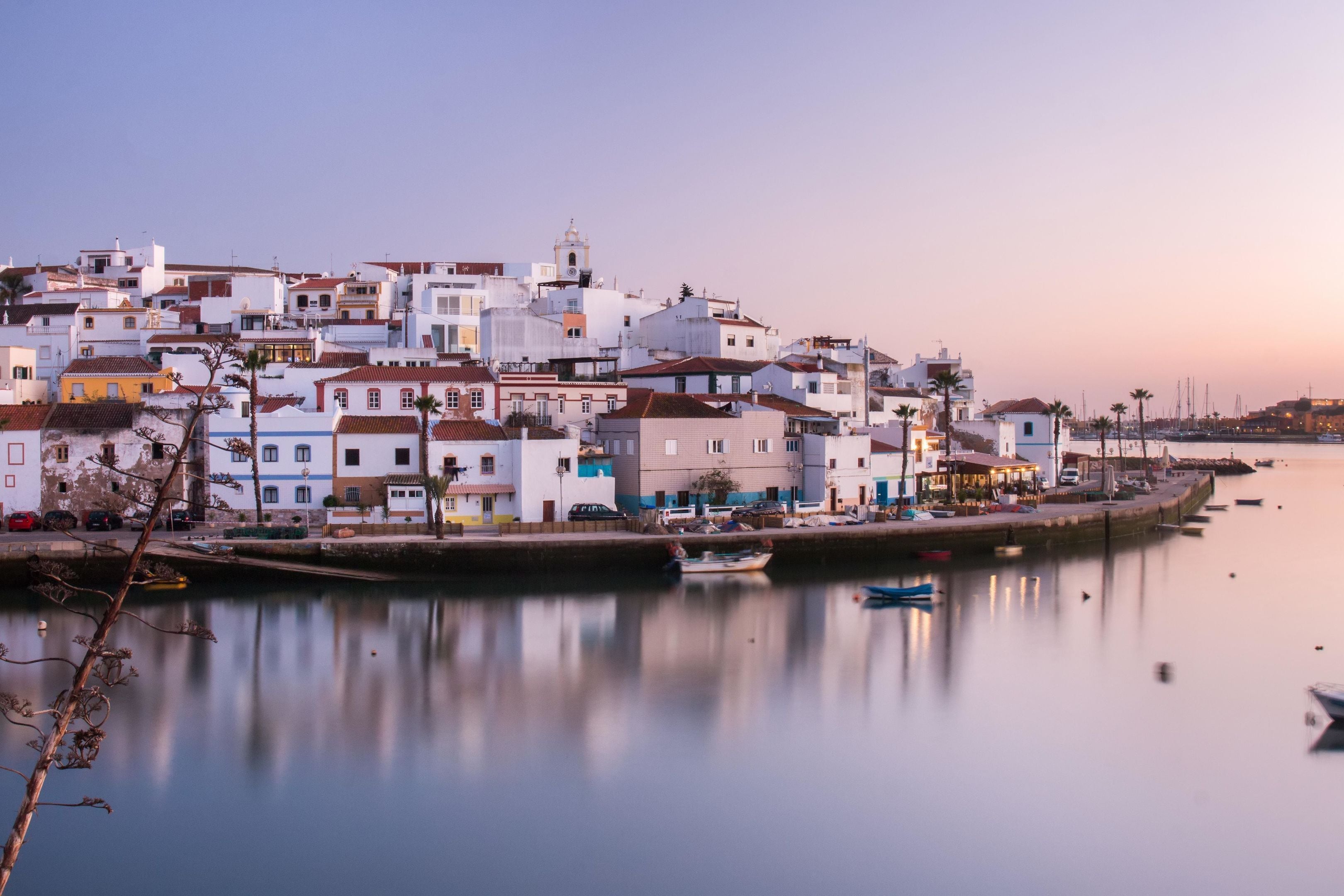 A coastal town with white buildings, a calm harbor, and a clear sky.