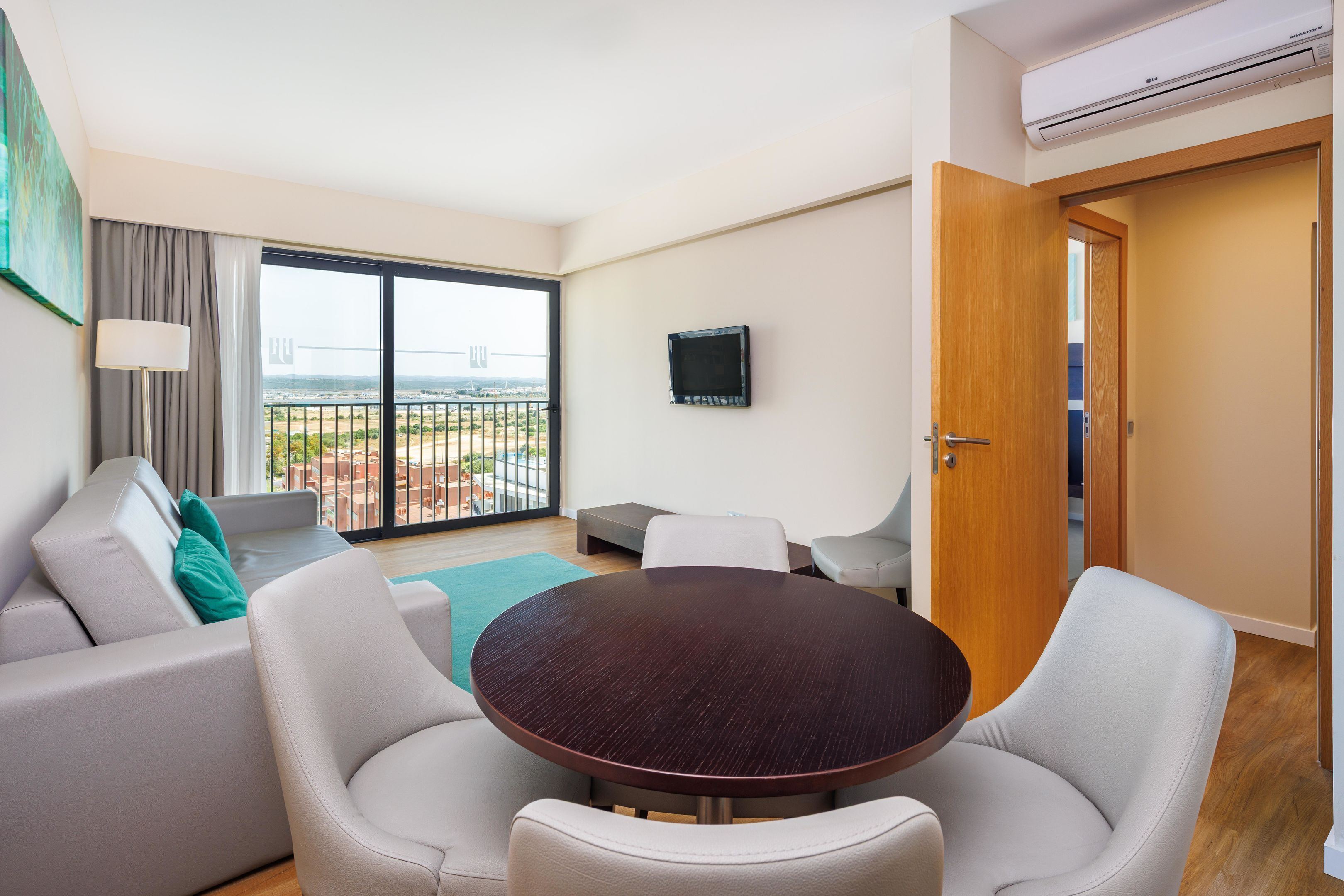 Een moderne woonkamer met een ronde tafel, witte leren banken, een aan de muur bevestigde televisie en een balkon met uitzicht op de stad.
