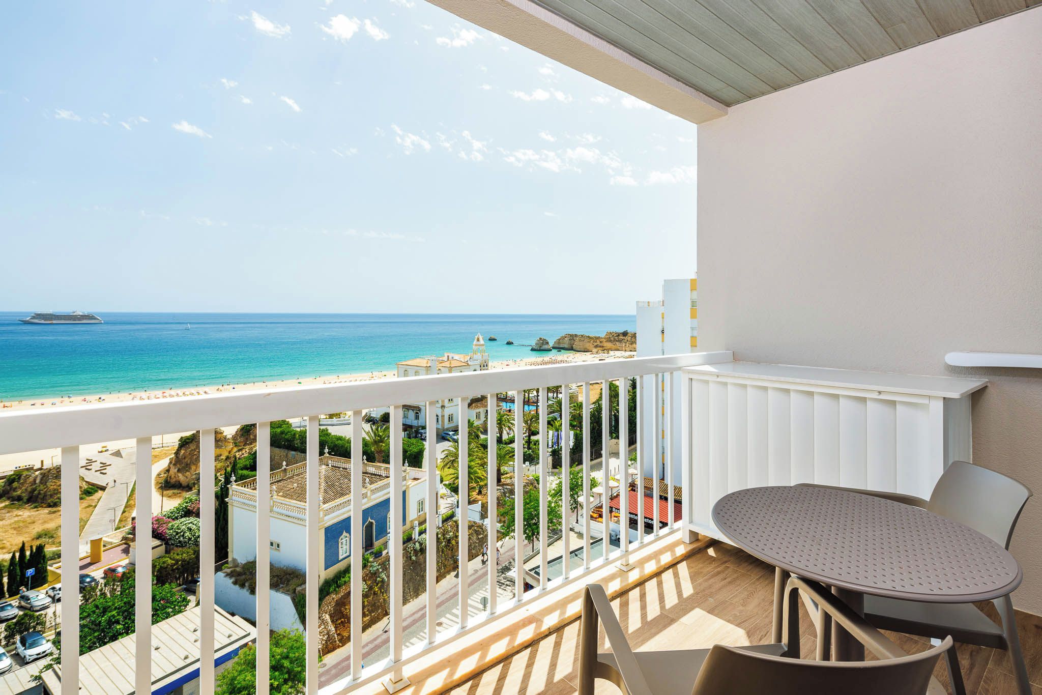 Un balcón con vista a la playa y al océano, una mesa con sillas y una barandilla blanca.