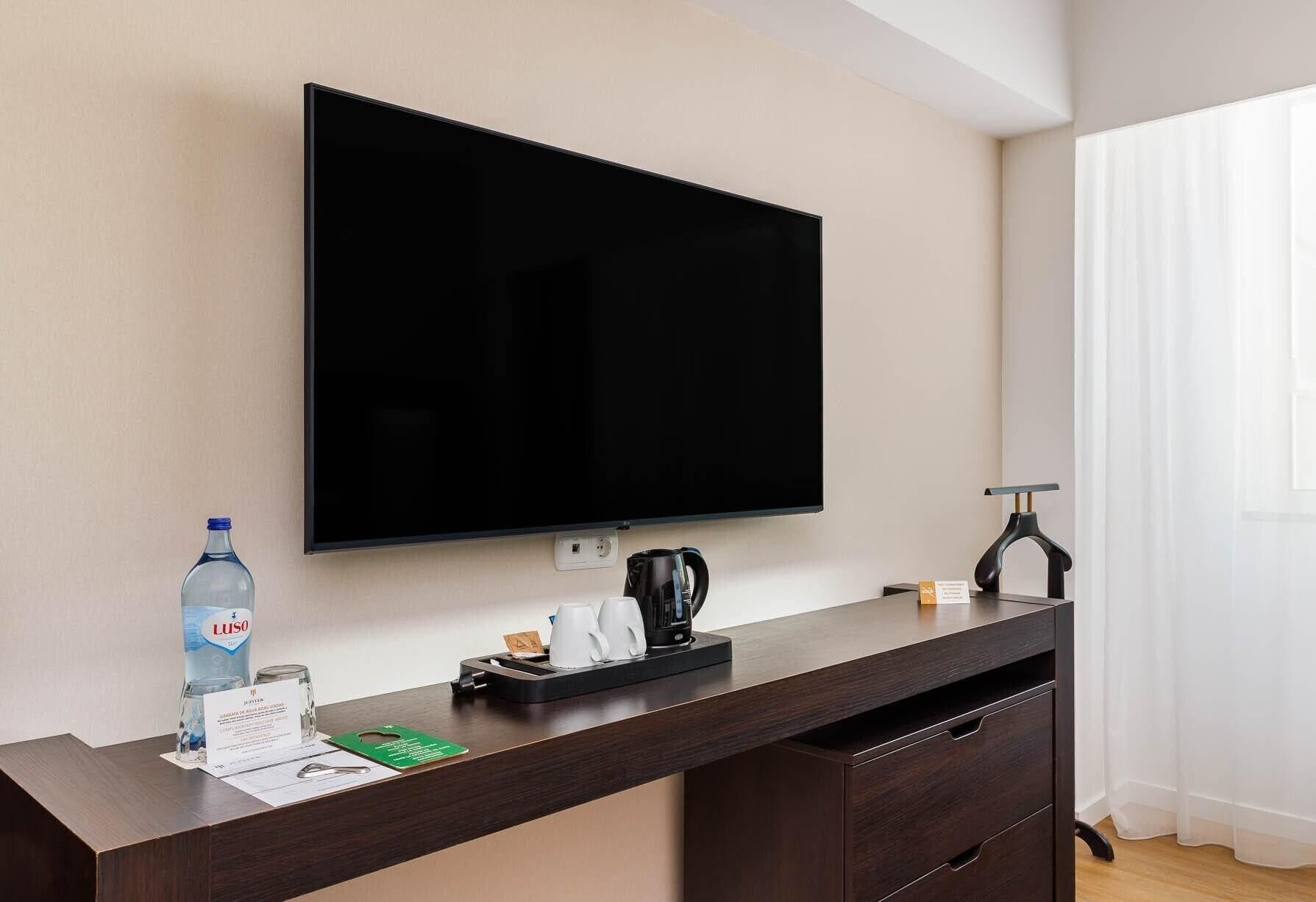 Quarto de hotel com televisão de tela plana na parede, cafeteira, uma garrafa de água e uma pequena mesa com etiqueta de nome.