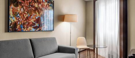 Une chambre d’hôtel moderne avec un canapé gris, une petite table ronde, une lampe sur pied et un grand tableau abstrait accroché au mur.