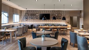 A modern dining area with round tables and chairs, a long counter with seating, and a large flat-screen TV mounted on the wall.