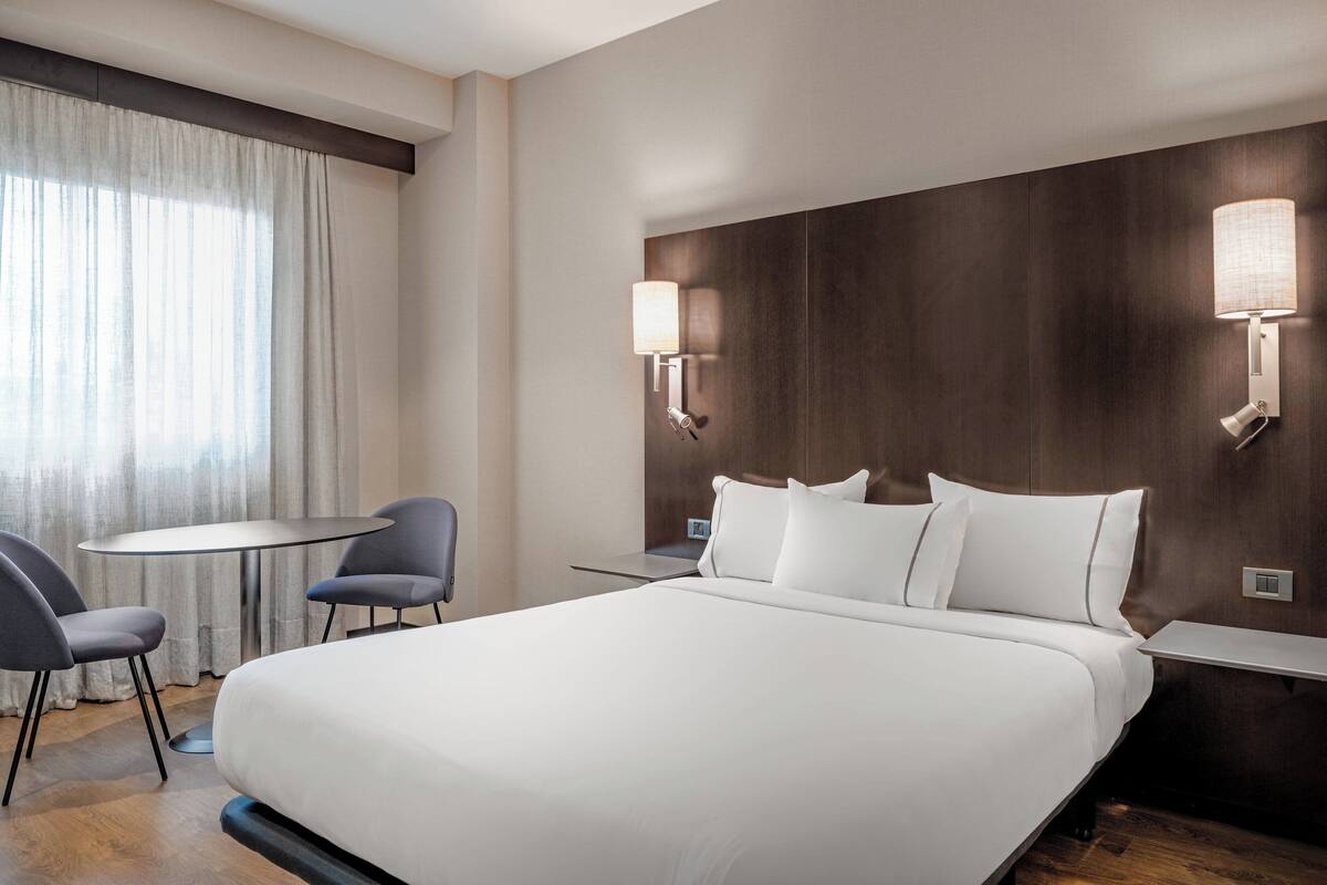 Une chambre d’hôtel avec un grand lit, deux tables de chevet, une petite table ronde avec des chaises et une fenêtre avec des rideaux.