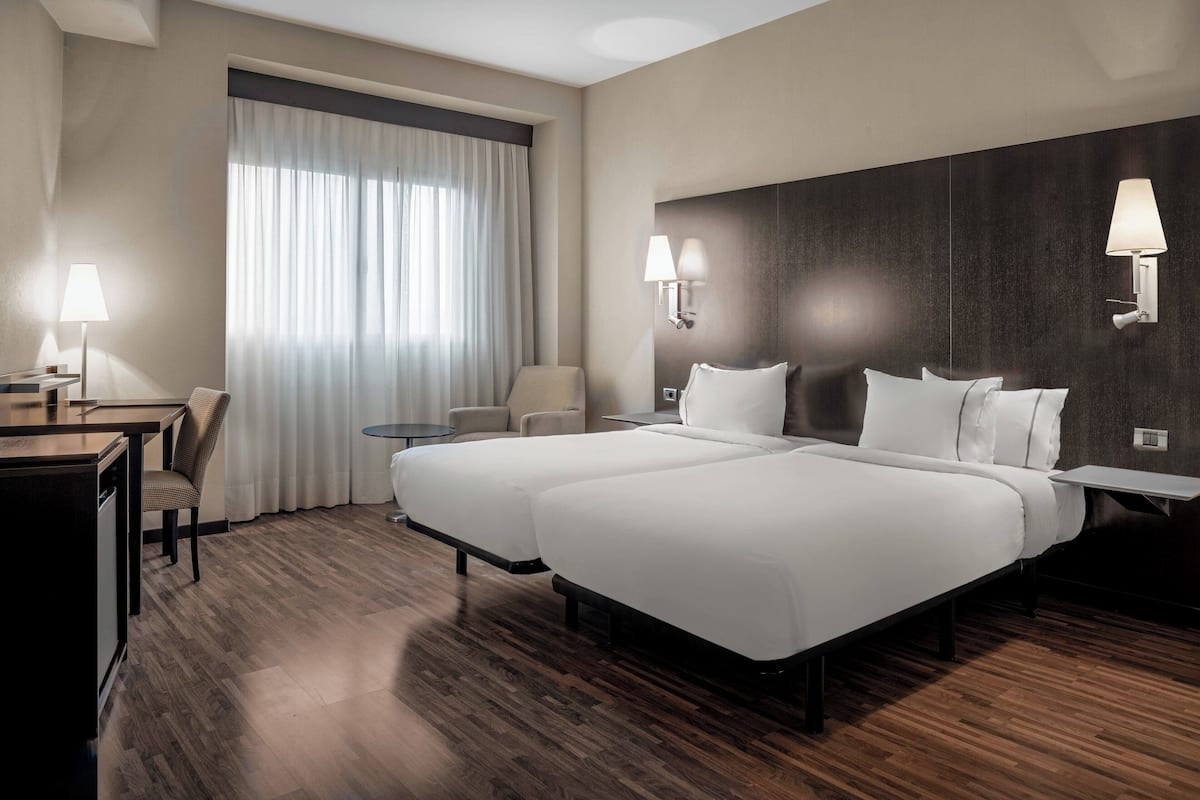 Une chambre d’hôtel avec deux lits, un bureau, une chaise et une petite table. Il y a deux lampes fixées au mur et une fenêtre avec des rideaux.