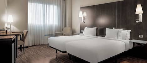 Habitación de hotel con dos camas, un escritorio, una silla y una mesita pequeña. Hay dos lámparas de pared y una ventana con cortinas.