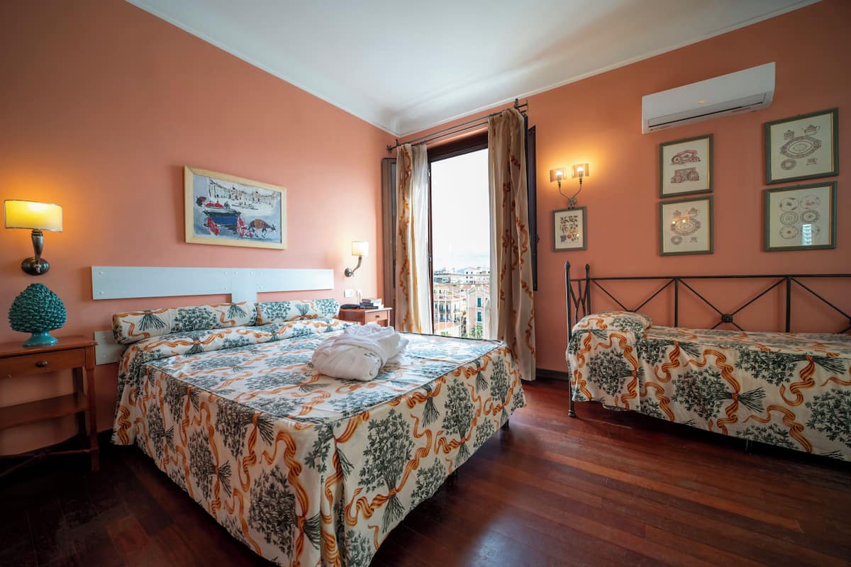 A hotel room with a bed, a bedside table, a lamp, a window with curtains, and wall art.