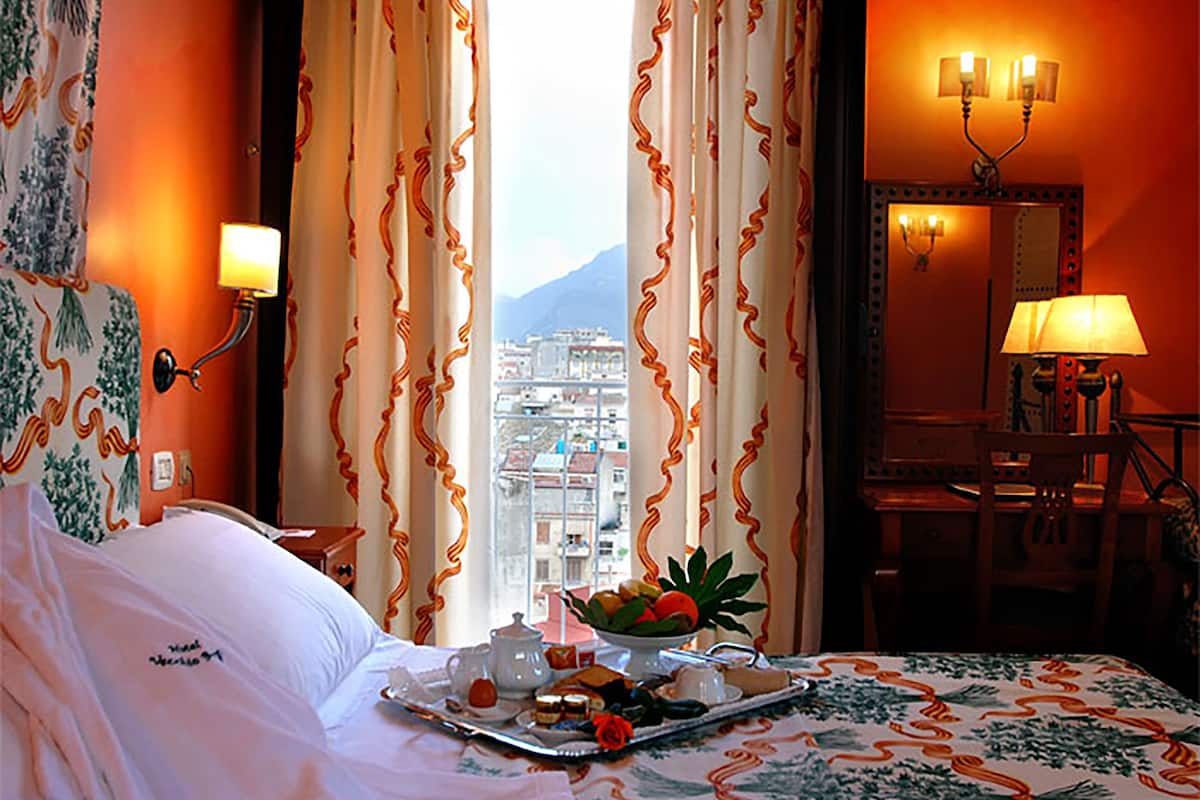 A hotel room with a bed, a tray of breakfast items, a view of a cityscape, and a desk with a lamp.