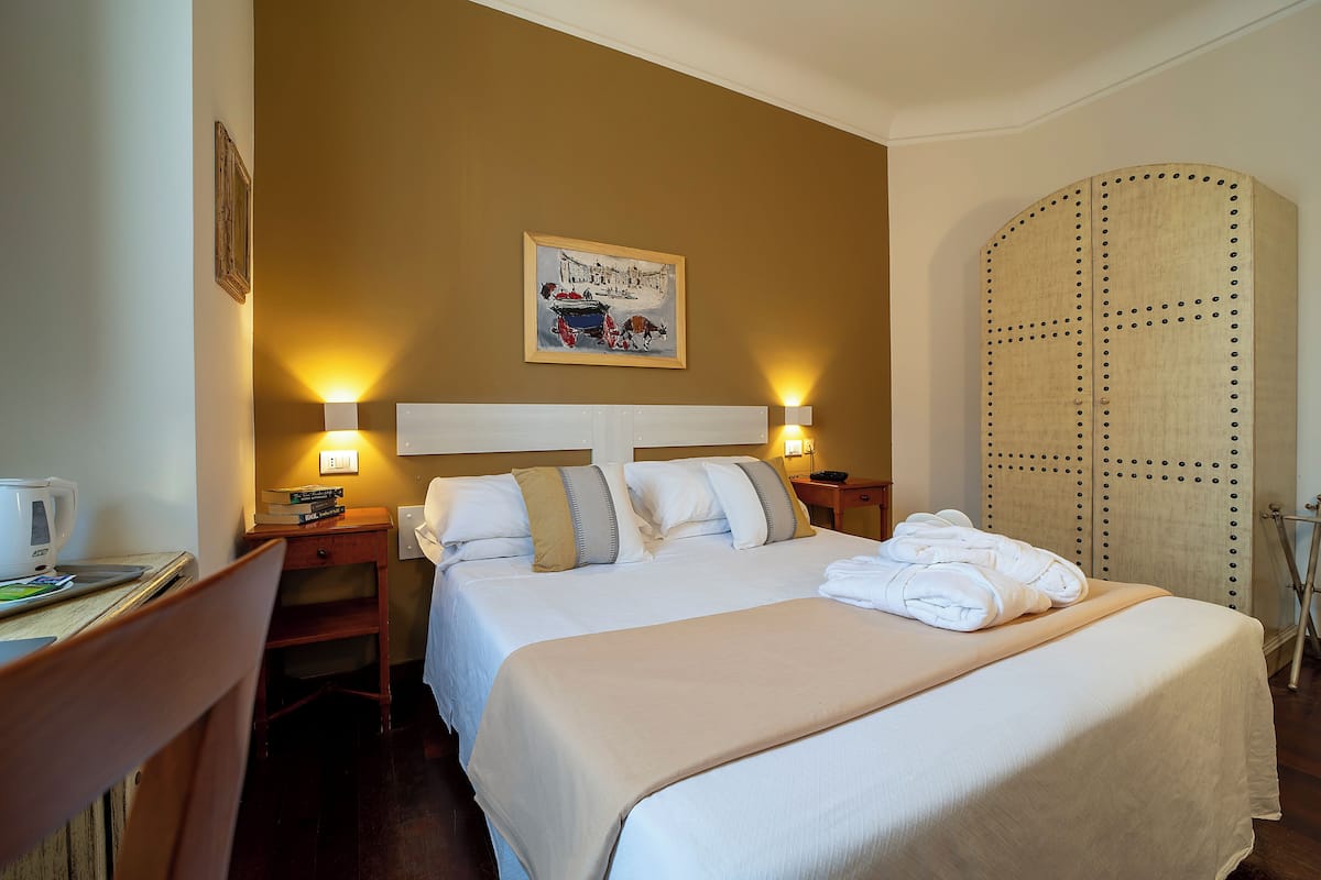 A hotel room with a bed, two bedside tables, a desk with a chair, a wall-mounted painting, and a wardrobe.
