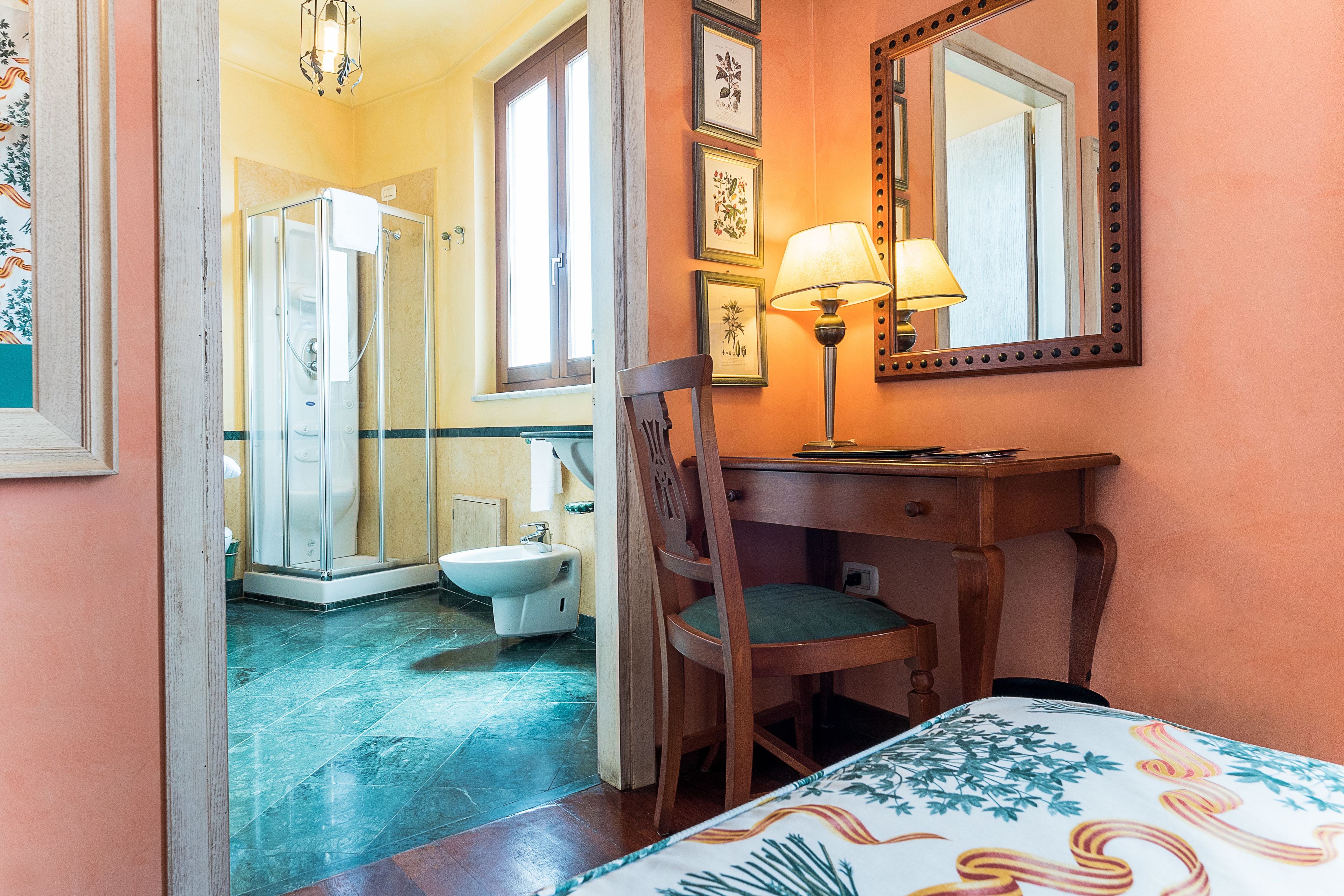 A bathroom with a shower, toilet, and sink. There is a chair and a table with a lamp.