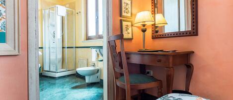 A bathroom with a shower, toilet, and sink. There is a chair and a table with a lamp.