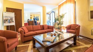 Un vestíbulo de hotel con un conjunto de sofá rojo, una mesa de centro de vidrio con libros y objetos decorativos, un mostrador de recepción y un espejo grande.