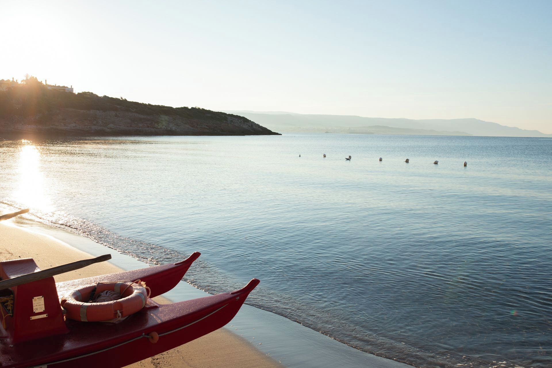 A calm sea with a boat docked on the shore, a lifebuoy, and a distant shoreline with hills.