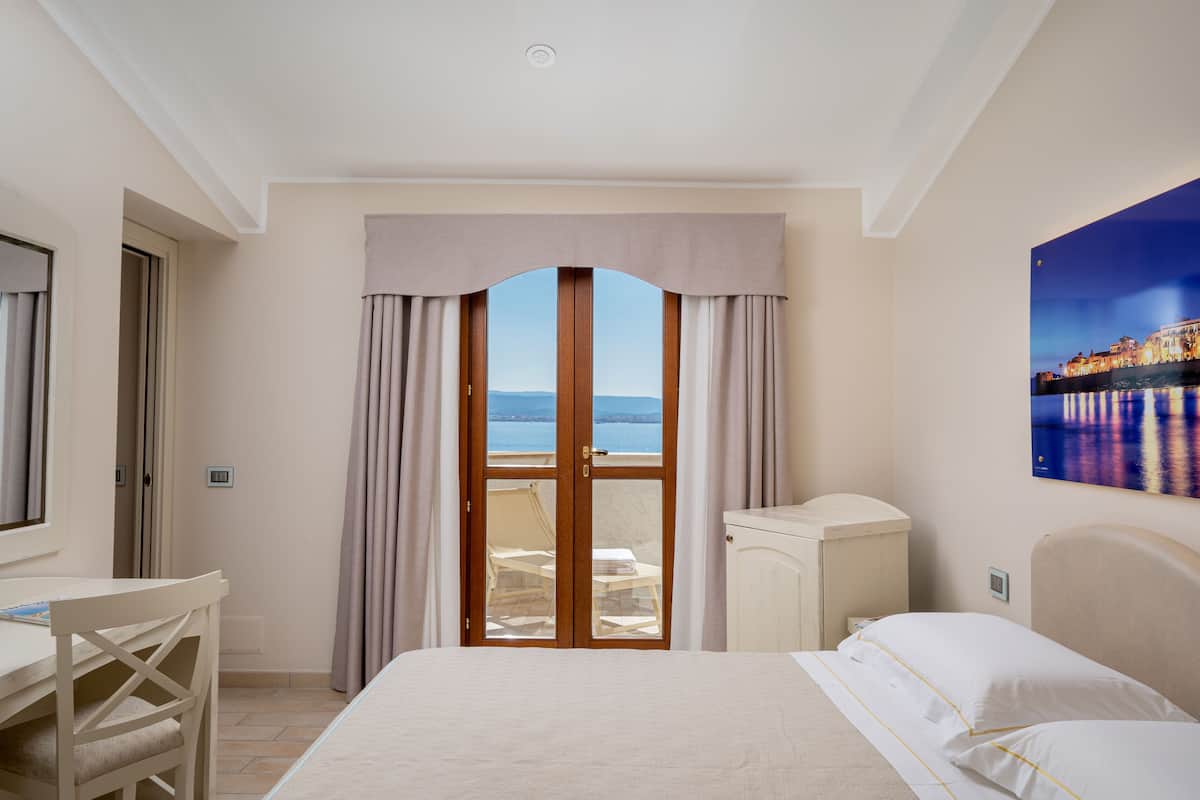 A hotel room with a bed, a desk, and a view of the sea through a glass door.