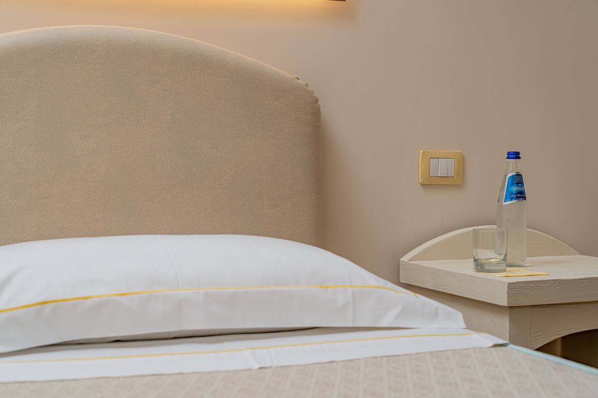 A neatly made bed with white bedding and a beige headboard. A small bedside table holds a water bottle and a glass.