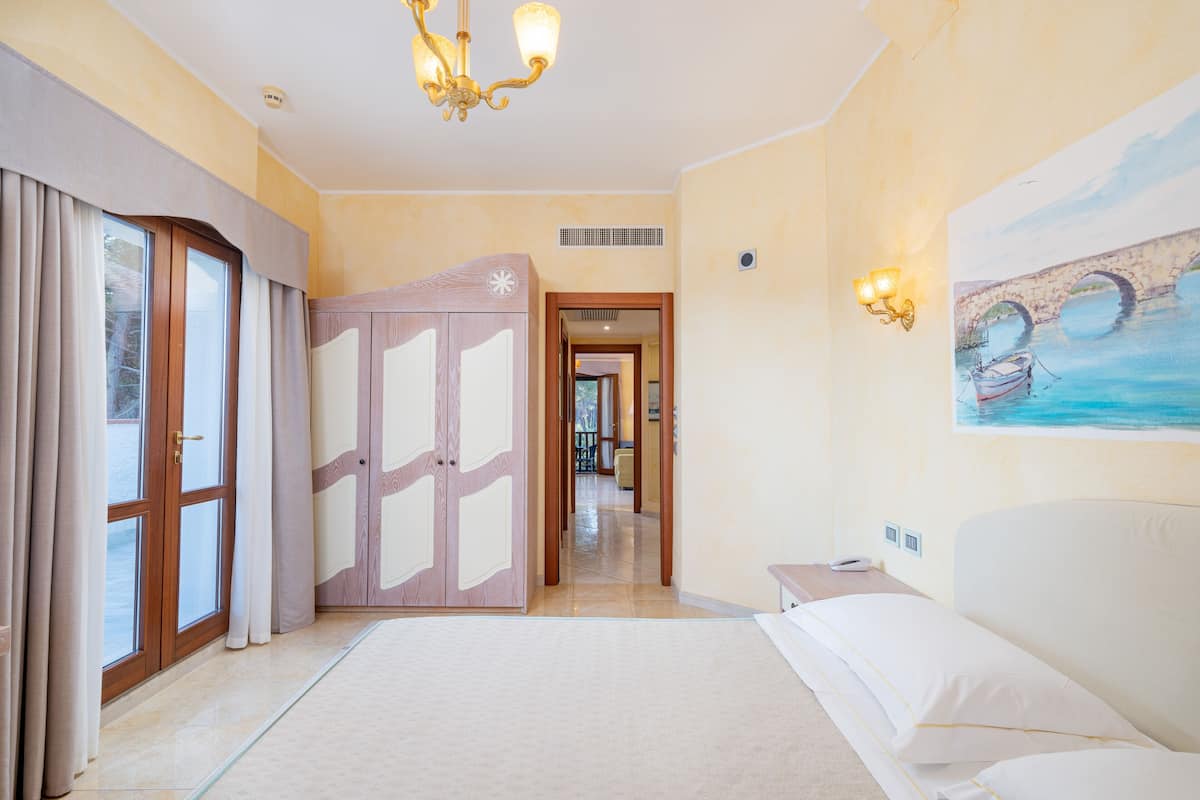 A hotel room with a large bed, a painting of a bridge, a chandelier, and a view of the outdoors through the window.