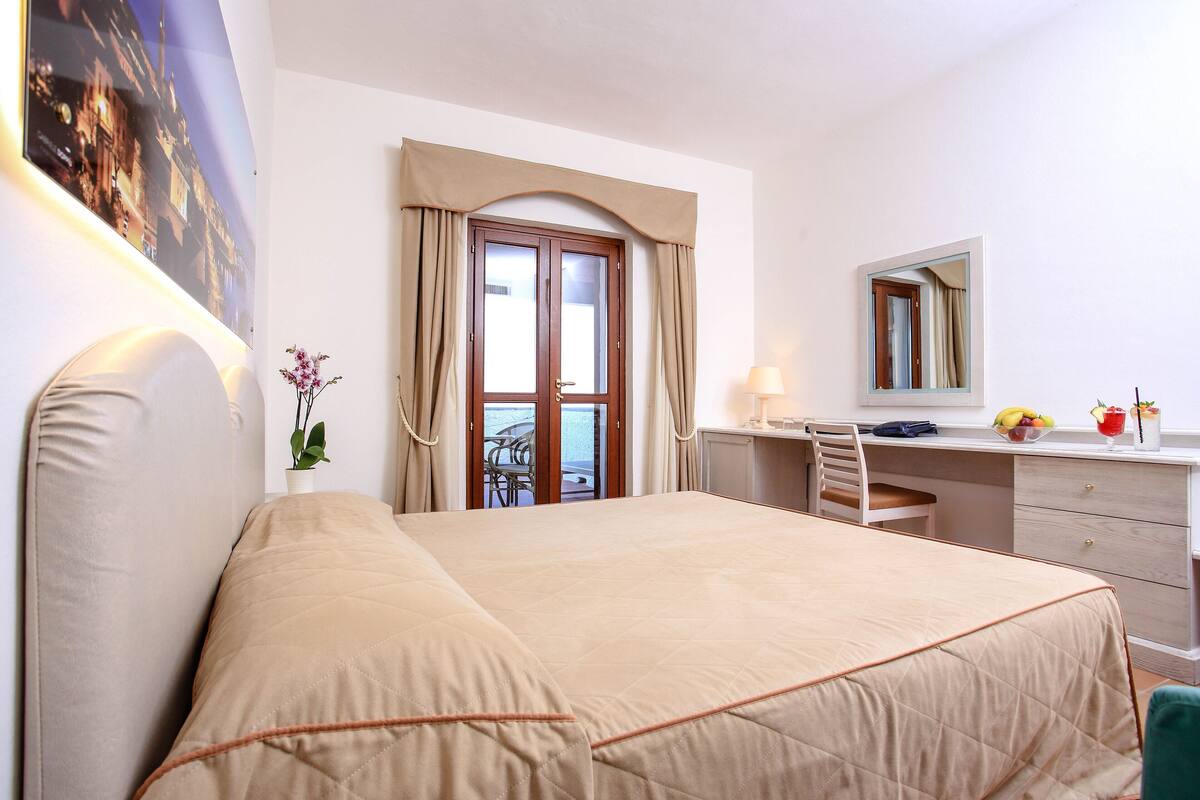 A hotel room with a bed, a desk, a chair, a mirror, a window with curtains, and a view of the outside.