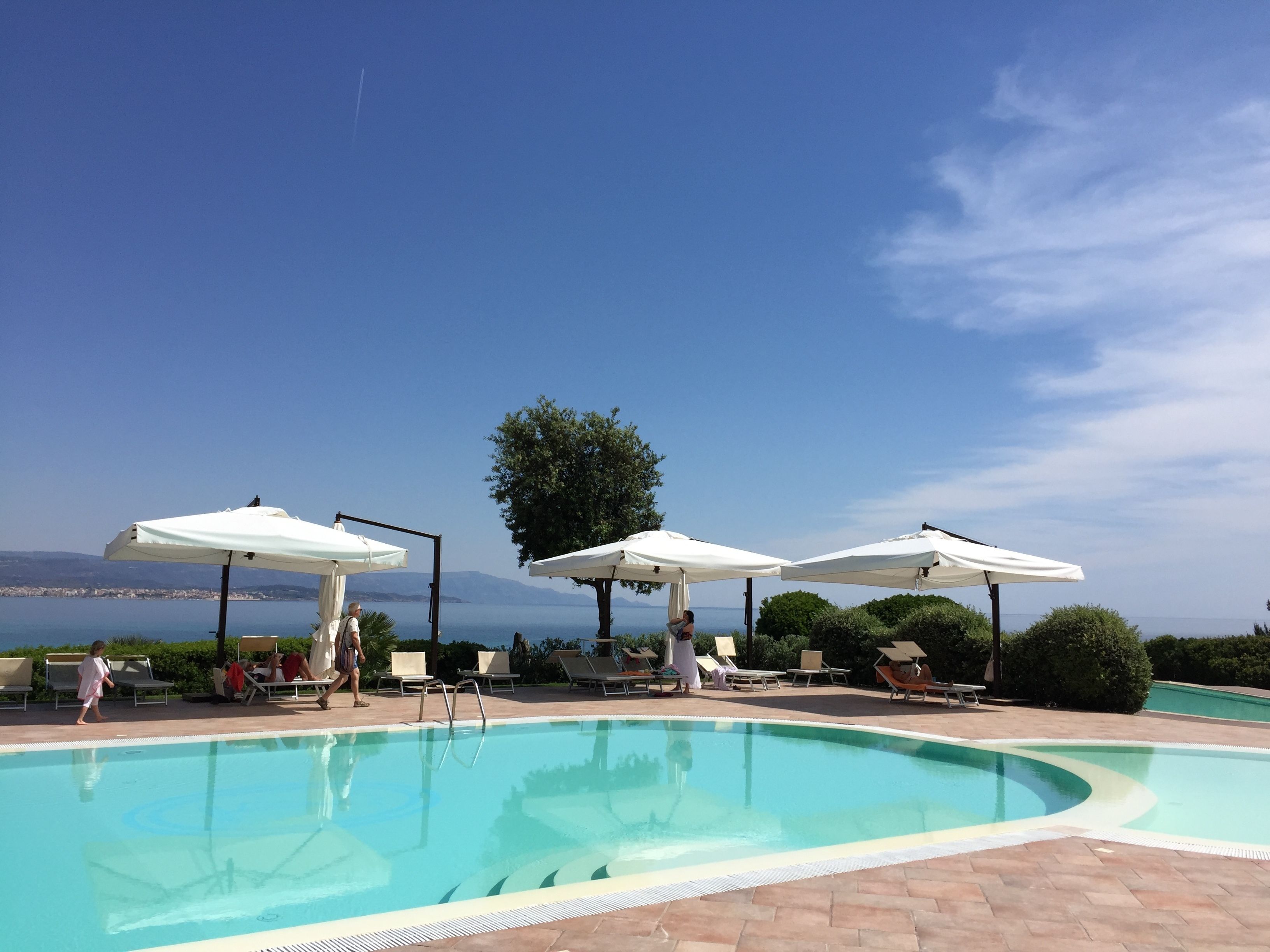 Piscina com espreguiçadeiras e guarda-sóis, cercada por vegetação e vista clara do mar.