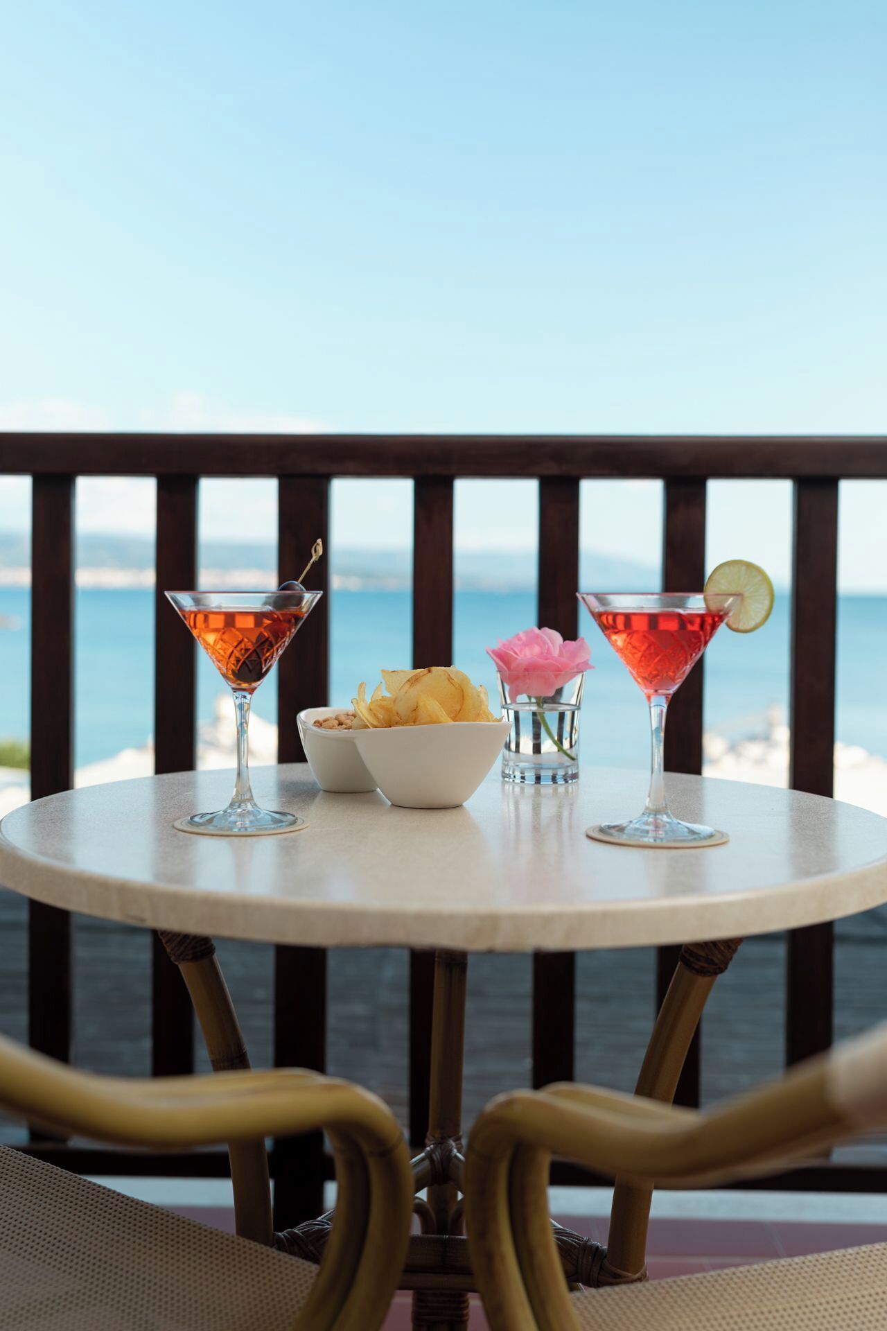 Une table avec deux verres à martini, un bol de chips et un verre d’eau, avec une rose, sur un balcon donnant sur la mer.