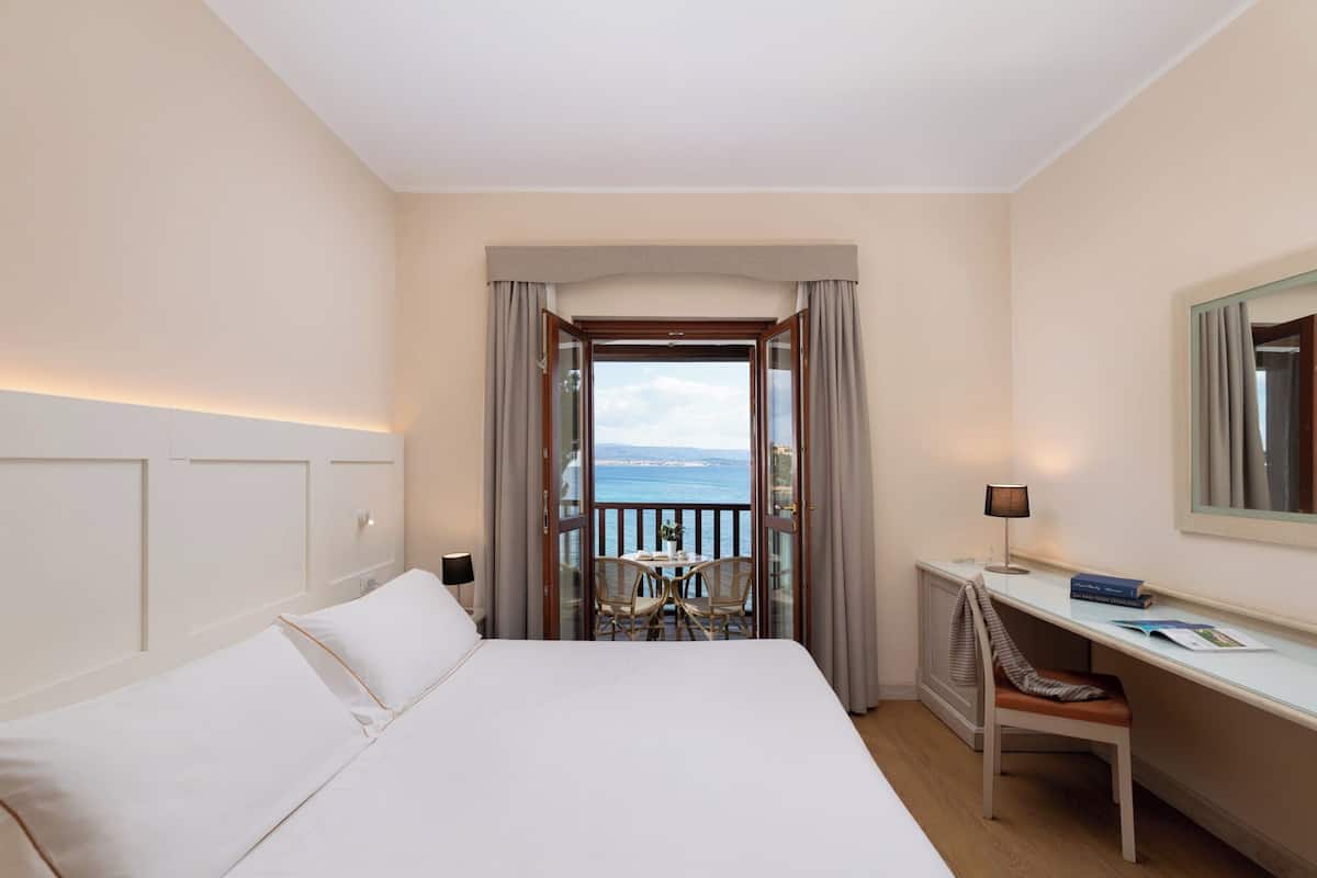A hotel room with a bed, a desk, a chair, and a view of the sea.