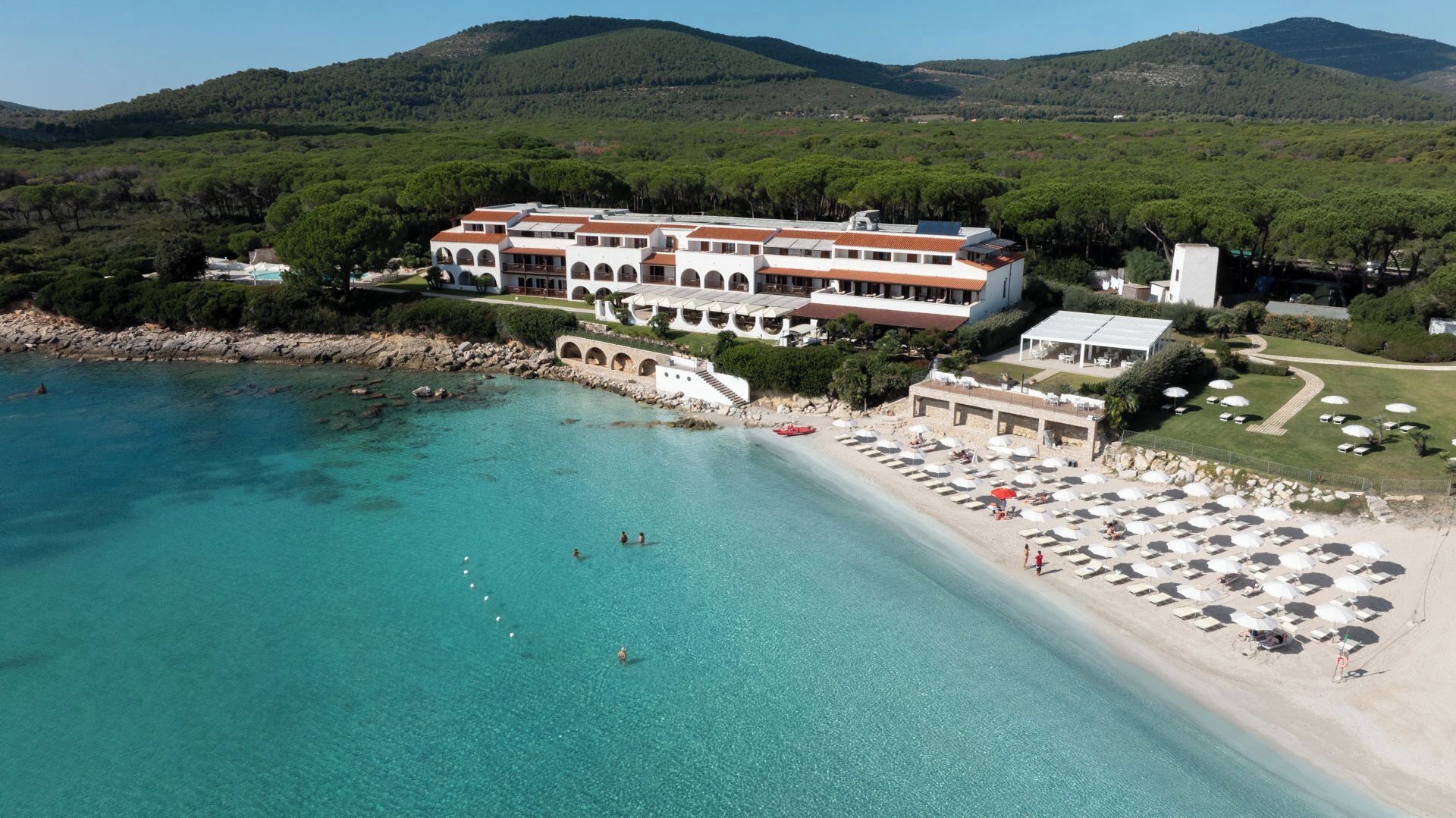 Un hôtel situé sur la côte, avec une plage, une eau cristalline et un paysage montagneux en arrière-plan.
