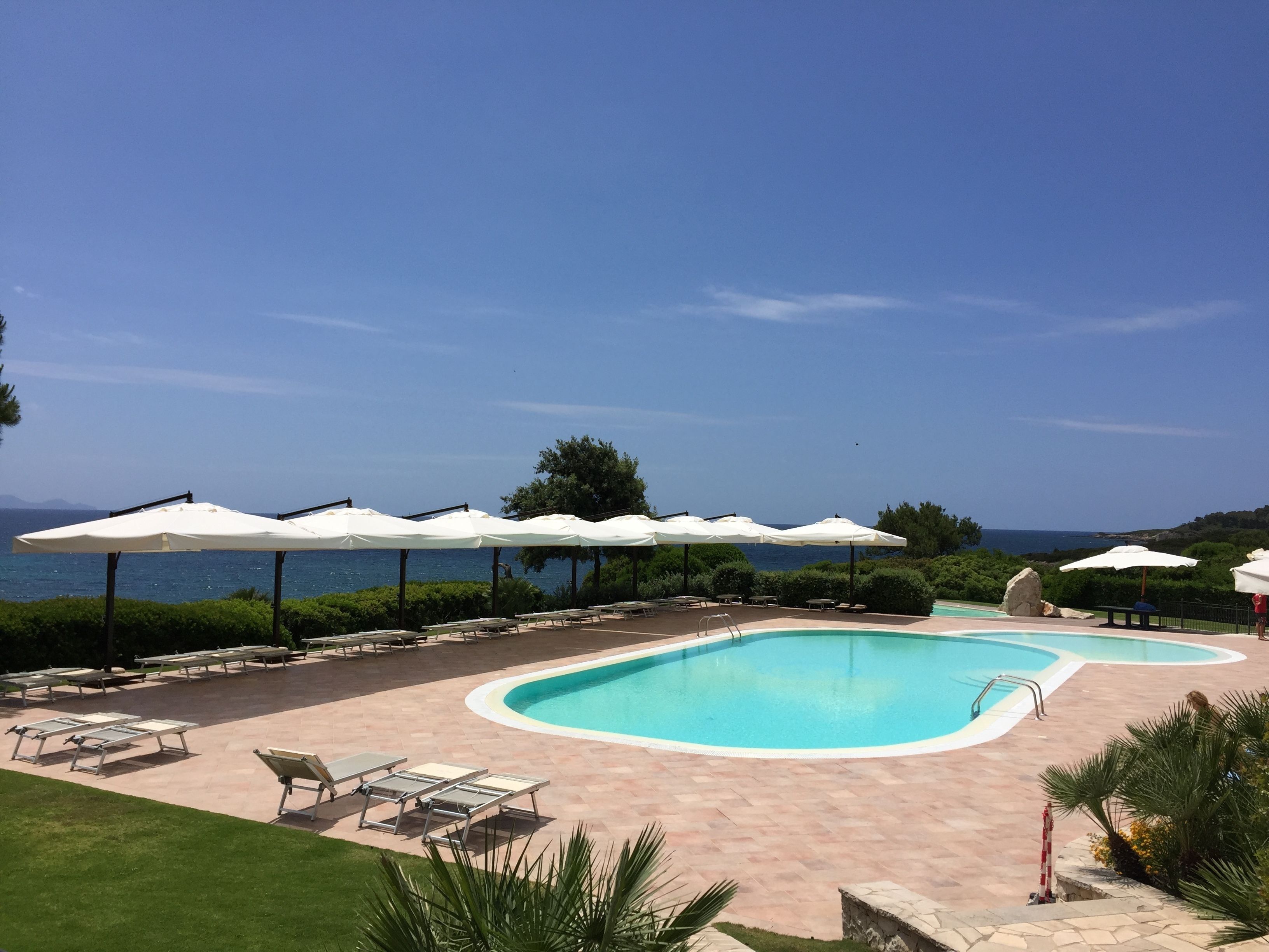 A pool area with lounge chairs and umbrellas, a clear sky, and a view of the sea.