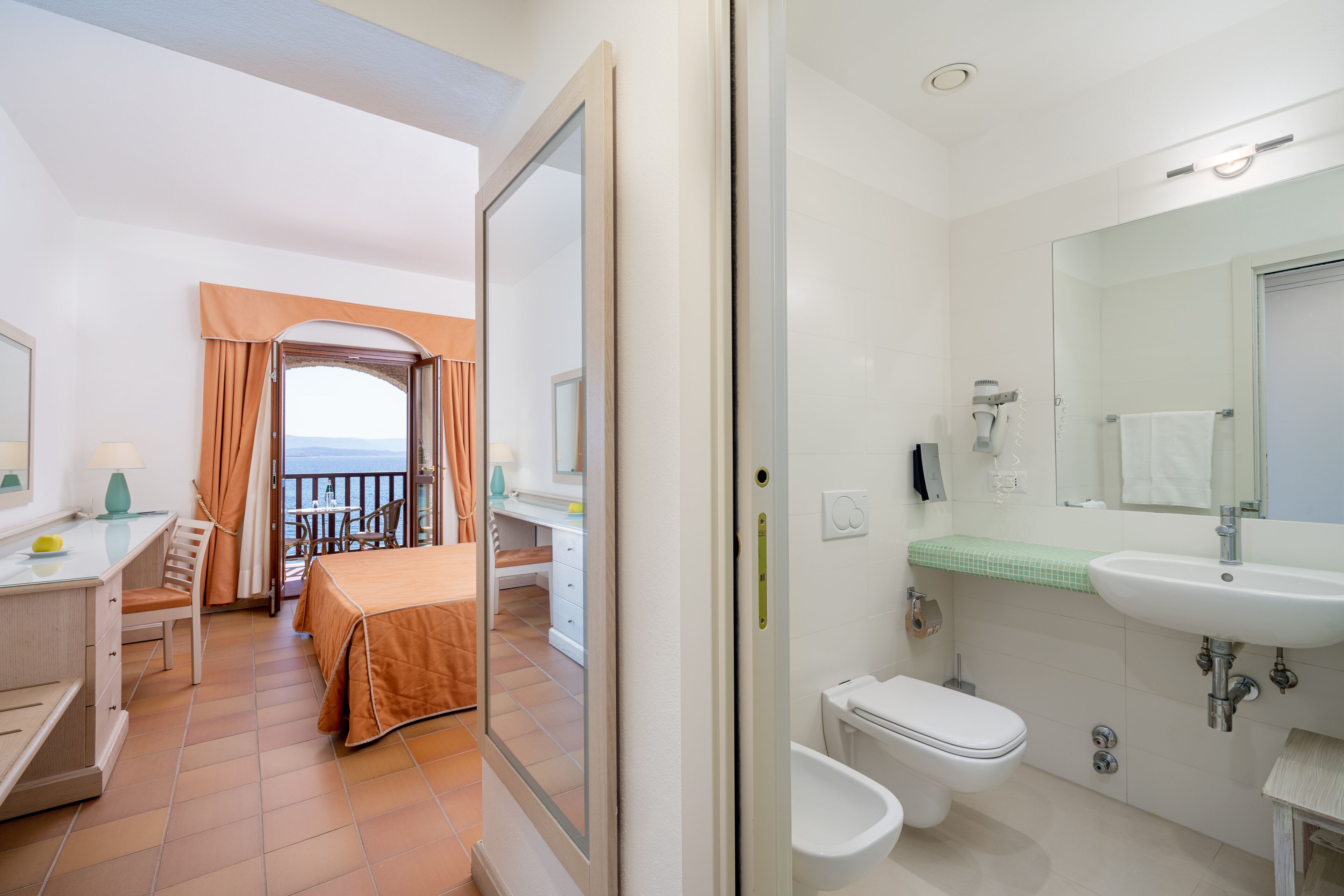 Habitación de hotel con balcón, escritorio con lámpara, cama con colcha naranja y baño con lavamanos y retrete.