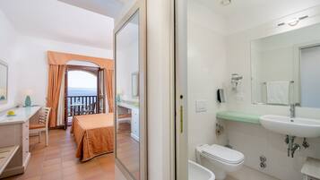 Habitación de hotel con balcón, escritorio con lámpara, cama con colcha naranja y baño con lavamanos y retrete.