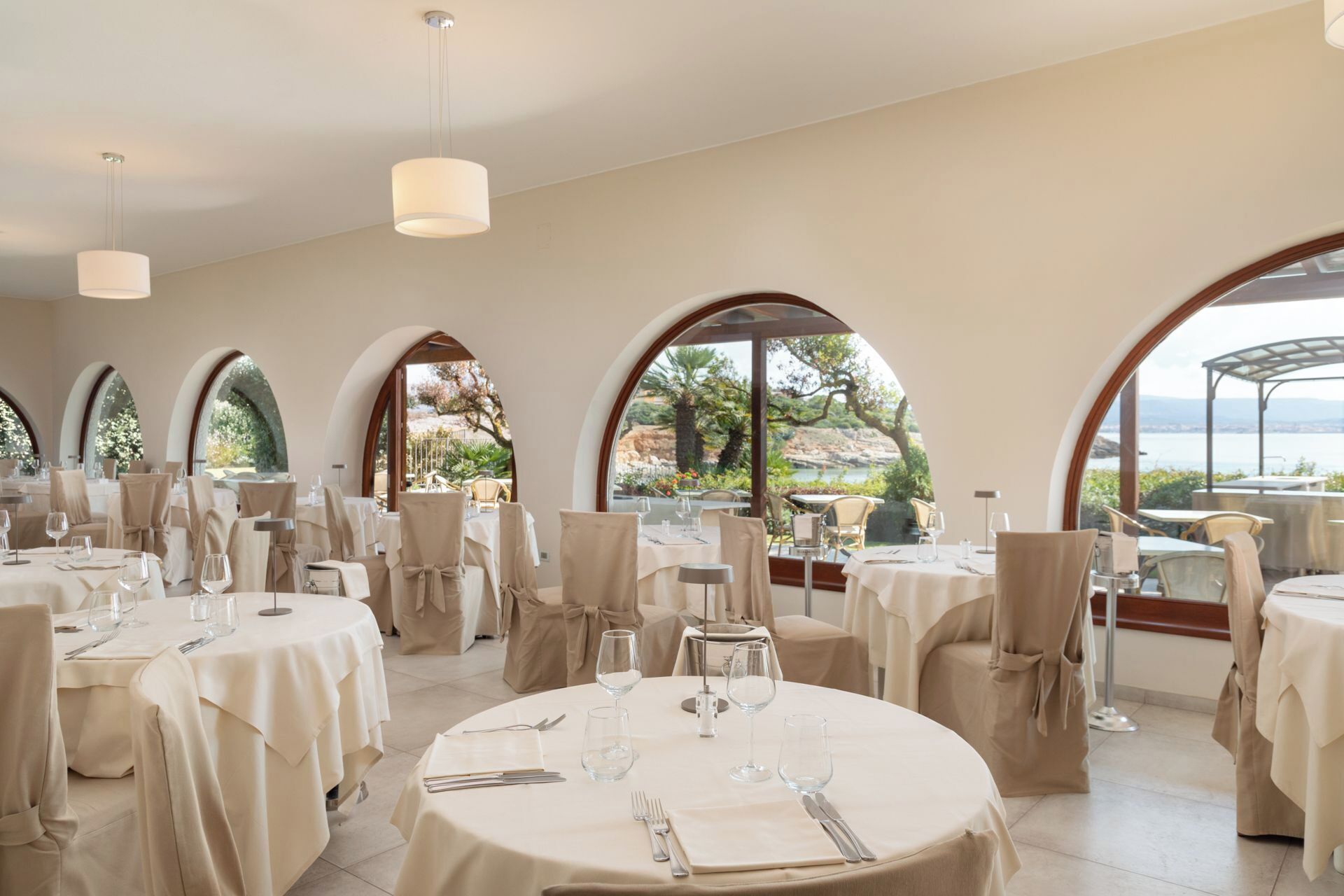 Een restaurant met ronde tafels, beige tafelkleden en stoelen met beige hoezen. Grote ramen met rondboogvorm en zicht op groen en een waterpartij.