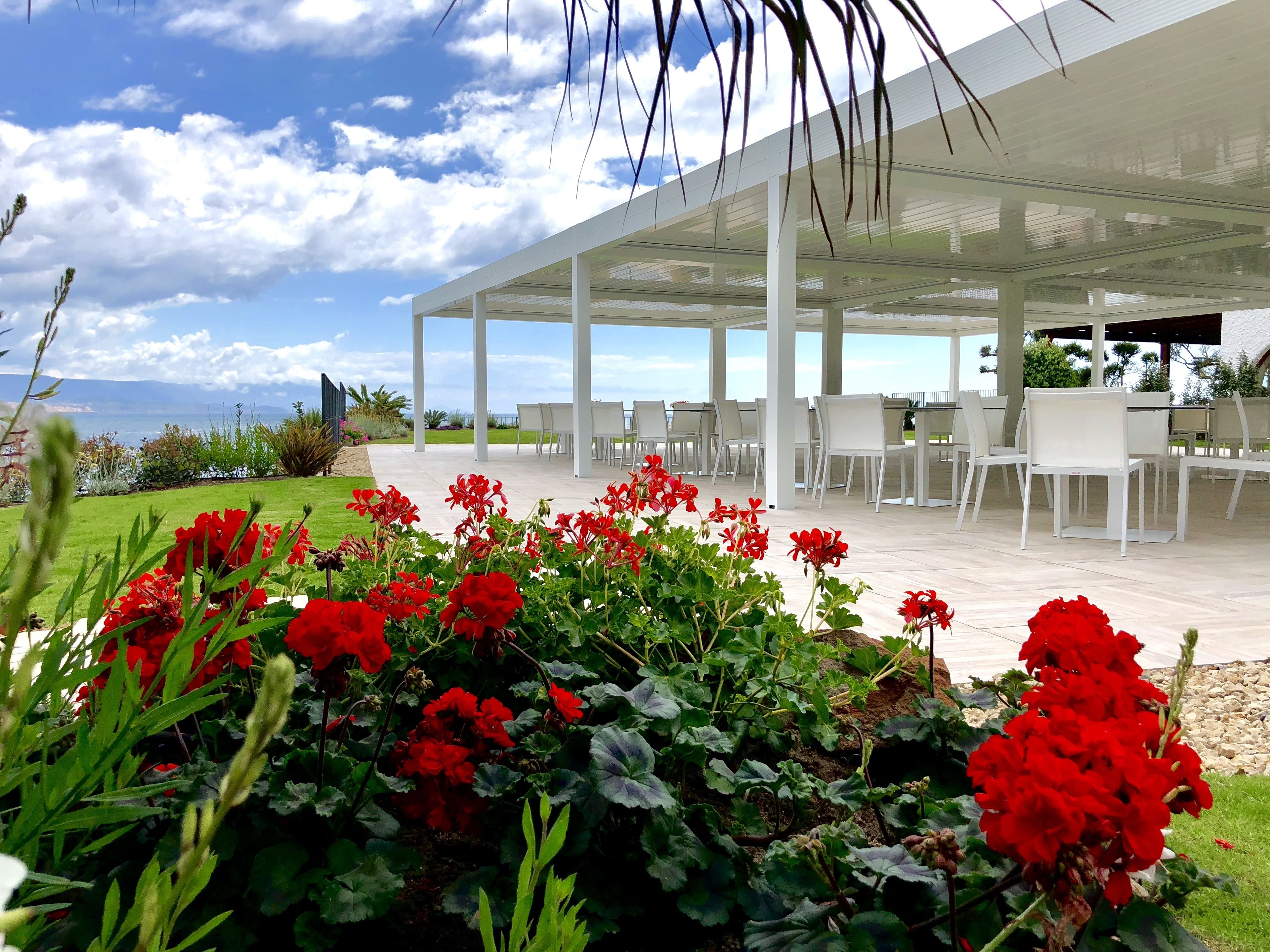 A covered outdoor seating area with white chairs and tables, surrounded by red flowers and greenery.