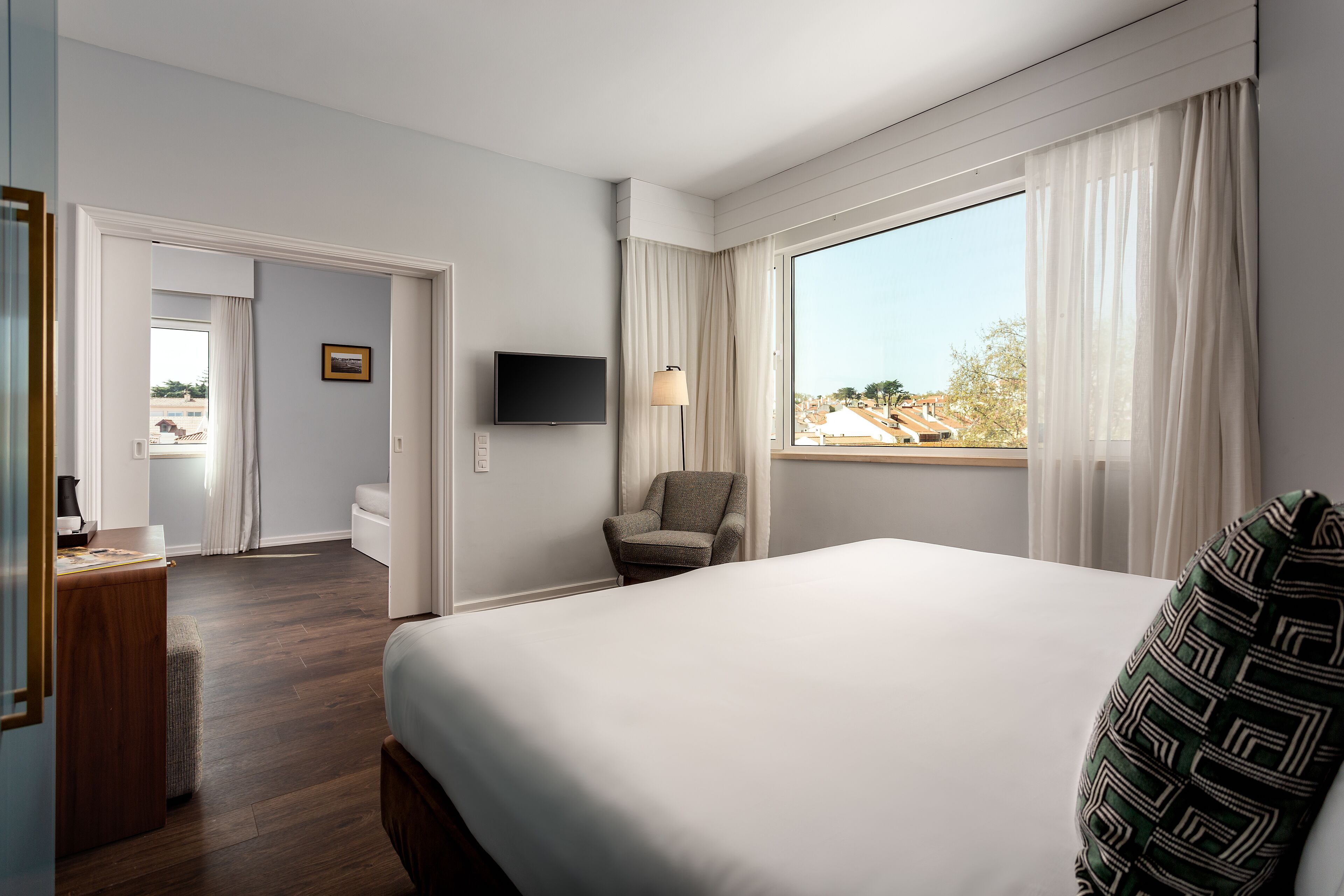 Ein Hotelzimmer mit einem großen Bett, einem Sessel, einem Schreibtisch, einem Fernseher und einem Fenster mit Vorhängen.