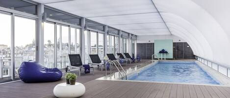 A modern indoor swimming pool with clear views of the city, lounge chairs, and a wooden deck.