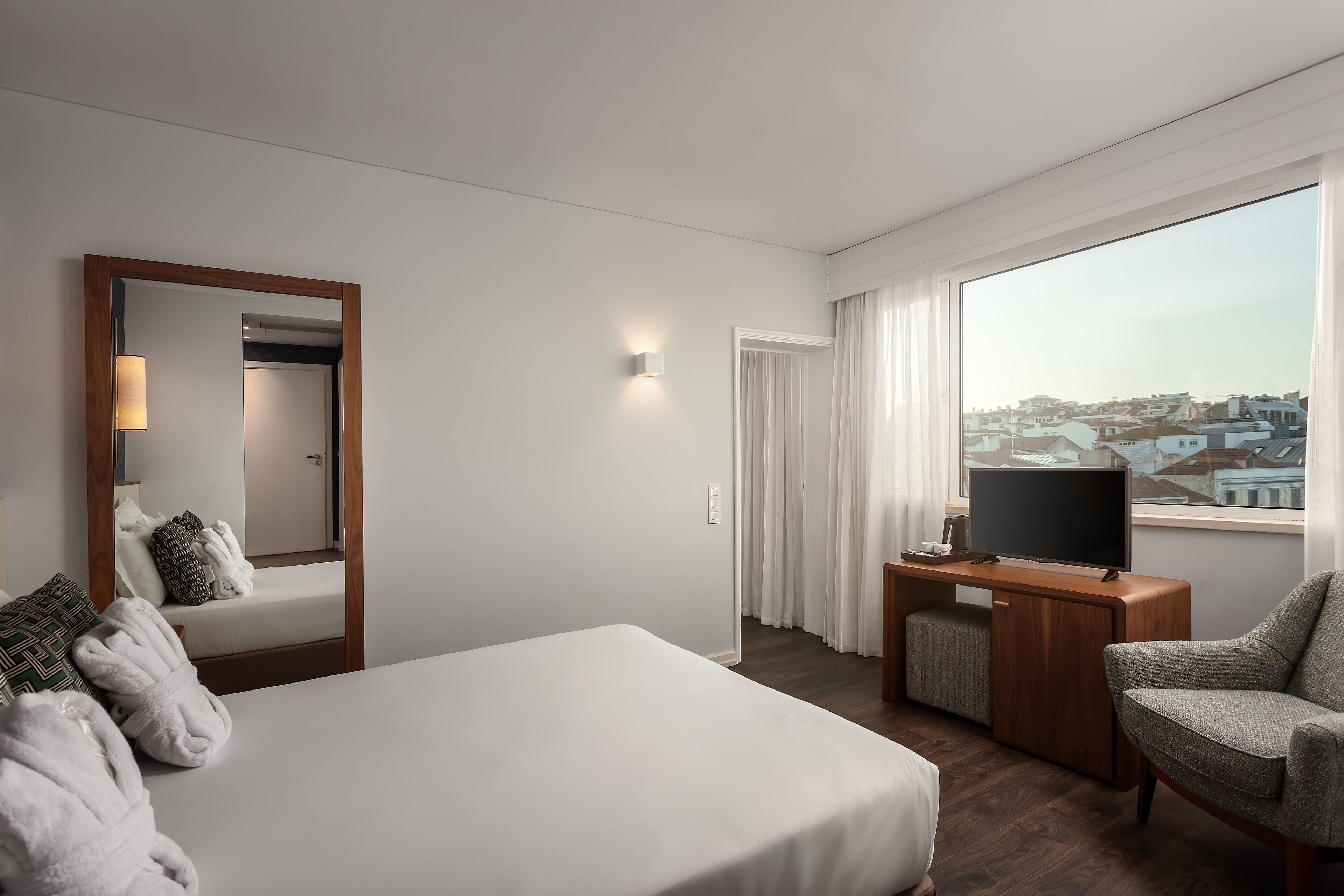 A hotel room with a bed, a TV on a wooden stand, a chair, and a large window with a city view.