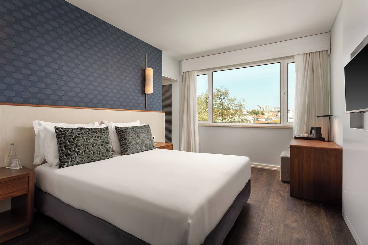 A modern hotel room with a large bed, a wooden nightstand, a desk, and a large window offering a city view.