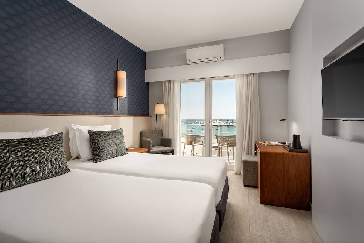 A hotel room with two beds, a desk, a chair, and a view of the sea.