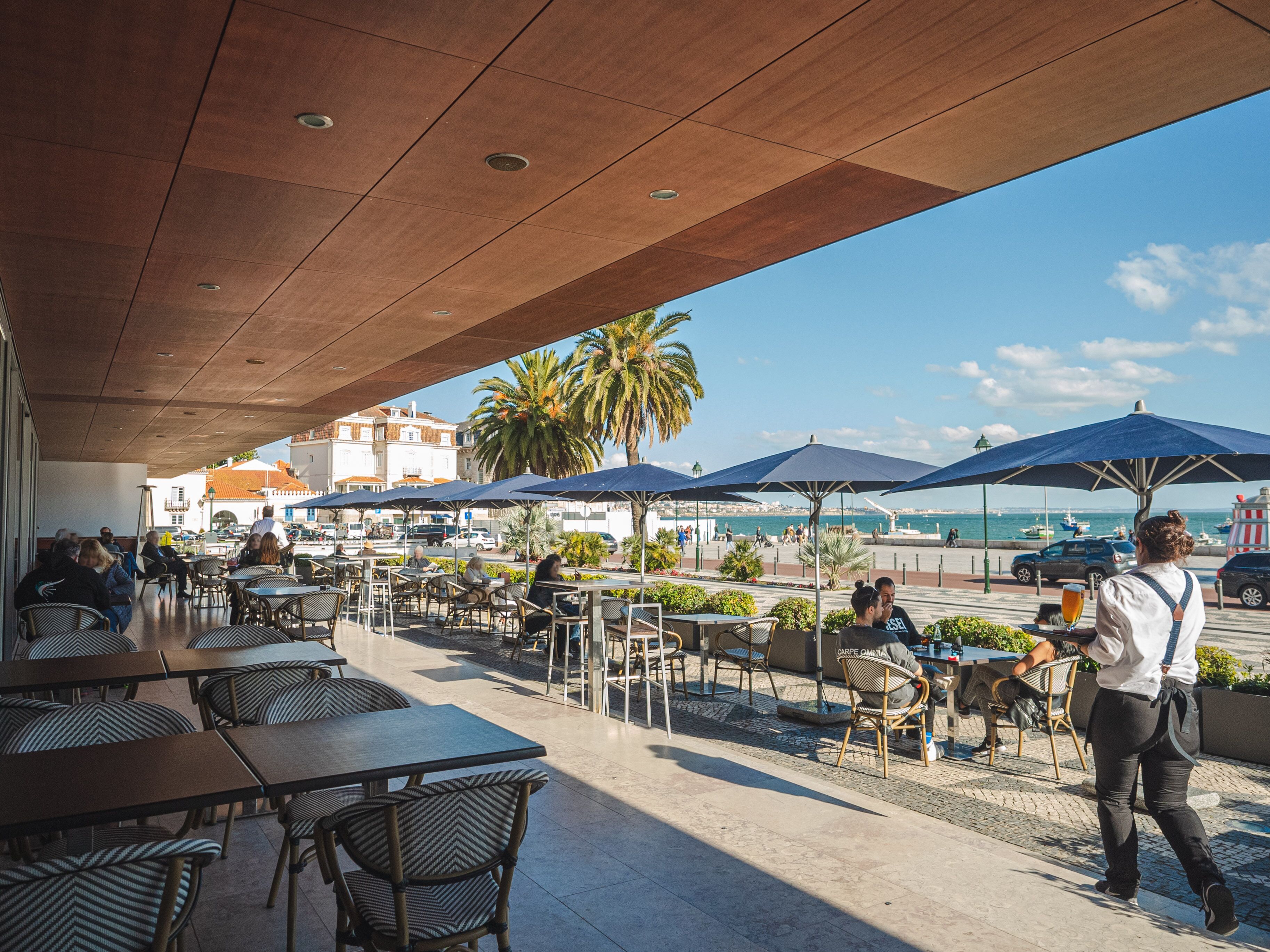 An outdoor café with tables and chairs, umbrellas, and a view of the sea.