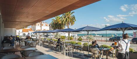 An outdoor café with tables and chairs, umbrellas, and a view of the sea.