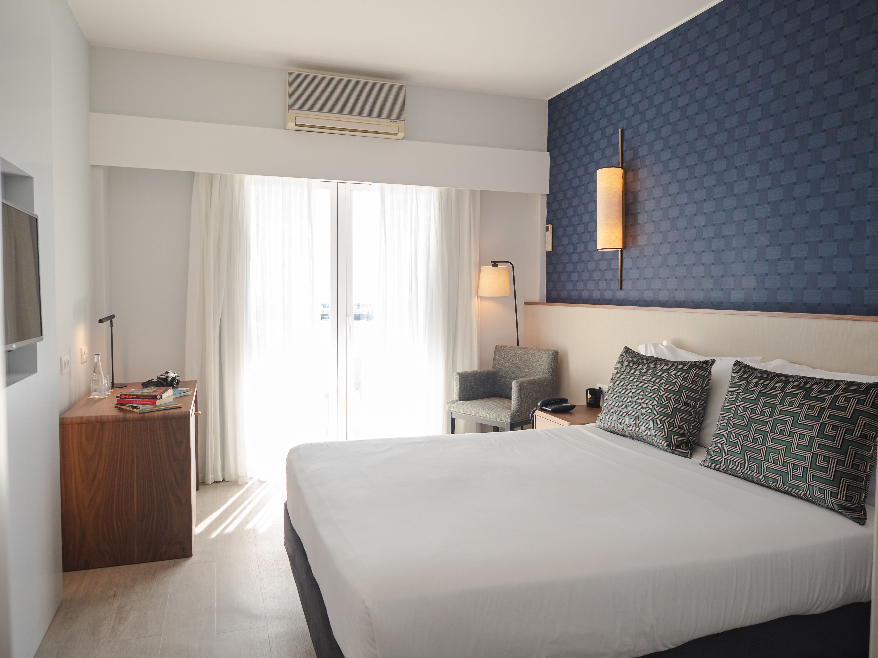 Une chambre d’hôtel avec un grand lit, un bureau avec une lampe, une chaise et un climatiseur fixé au mur.