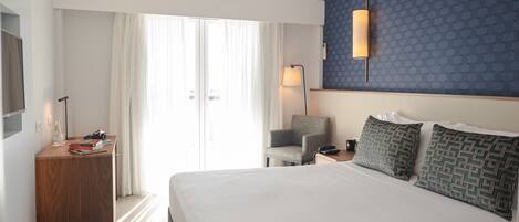 A hotel room with a large bed, a desk with a lamp, a chair, and a wall-mounted air conditioner.