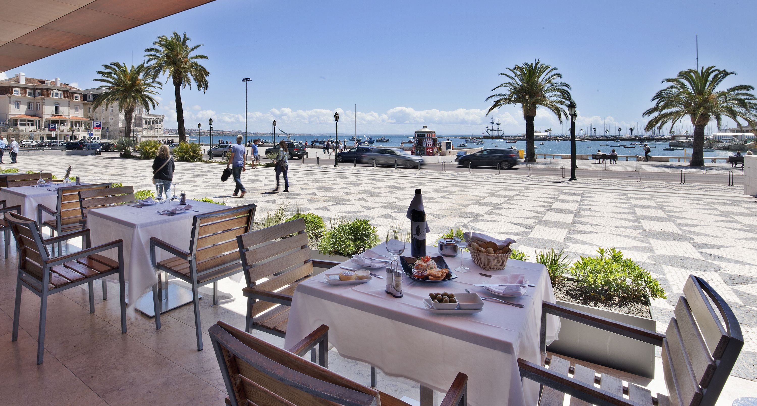 An outdoor café with tables and chairs, a view of the sea, and palm trees.