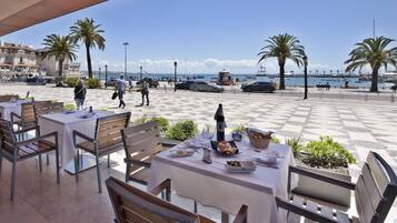 An outdoor café with tables and chairs, a view of the sea, and palm trees.