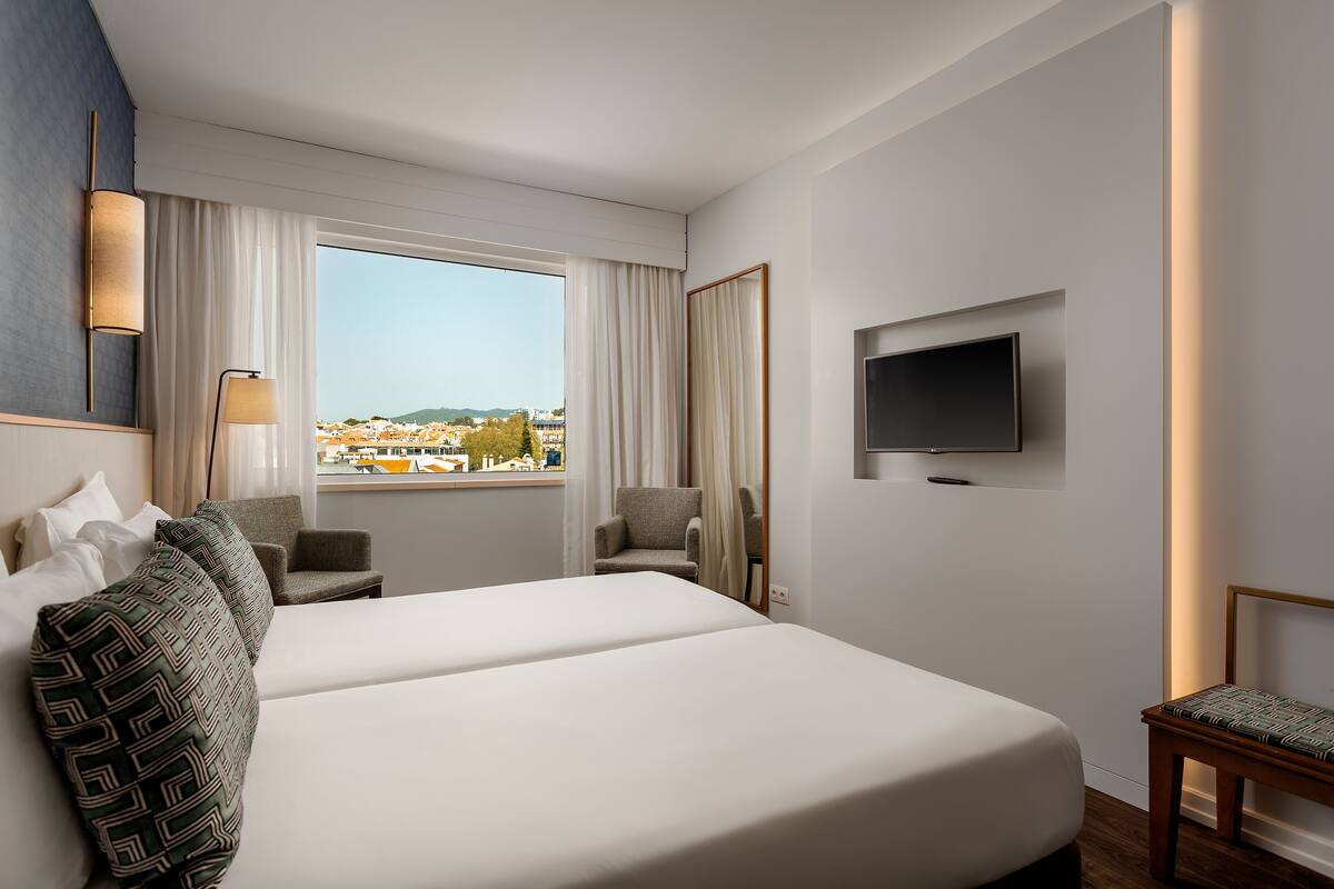 A hotel room with a large bed, a TV mounted on the wall, a chair, and a view of the city.