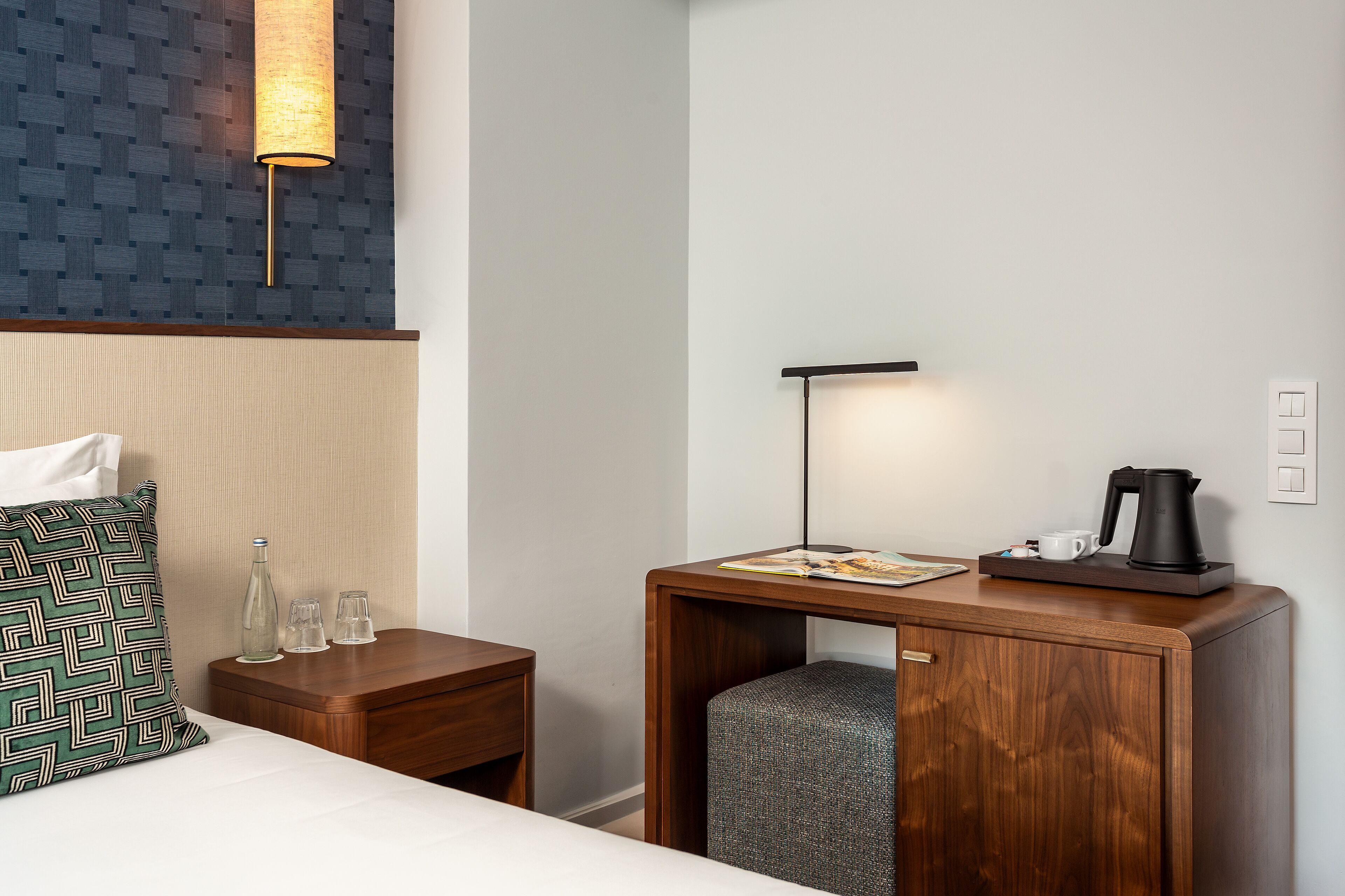 Quarto de hotel com cama, mesa de cabeceira com abajur, pequena mesa de madeira com chaleira, xícaras e revista.