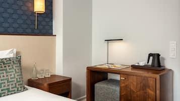 Quarto de hotel com cama, mesa de cabeceira com abajur, pequena mesa de madeira com chaleira, xícaras e revista.