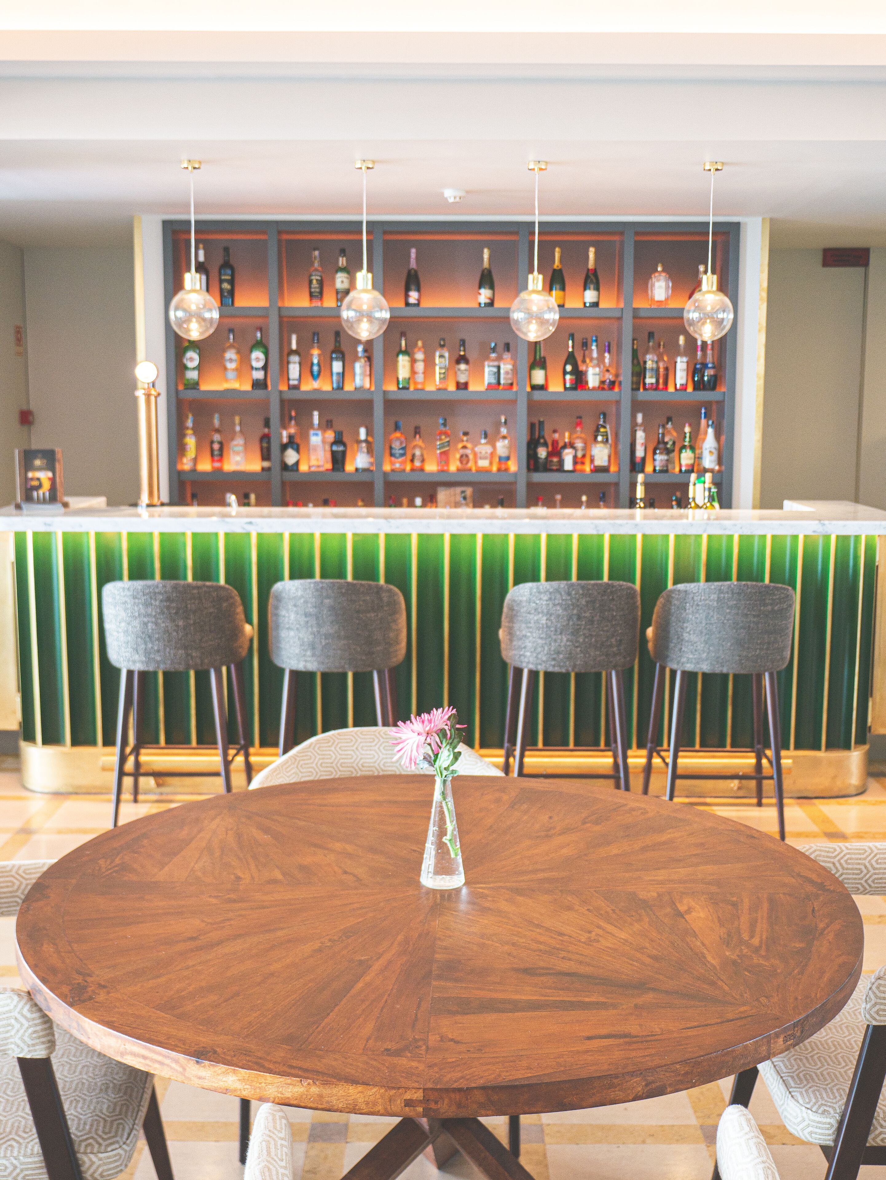 Una zona de bar con una barra verde, varias estanterías llenas de botellas de licor y tres taburetes.